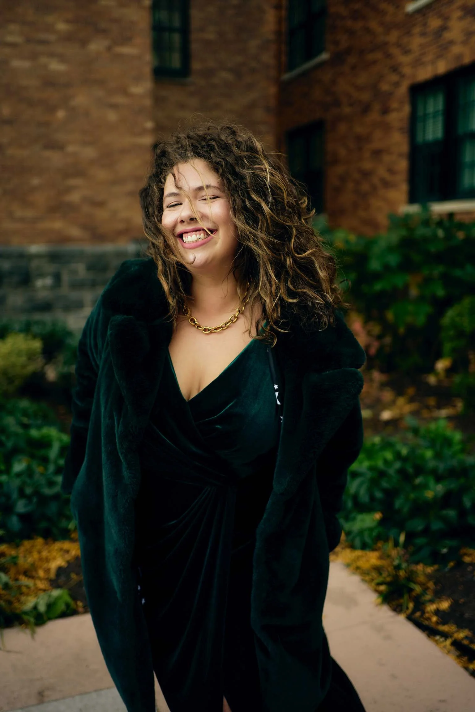 Bailey Jenkins smiling and posing outdoors in front of a brick building, wearing a black dress and a gold chain necklace.