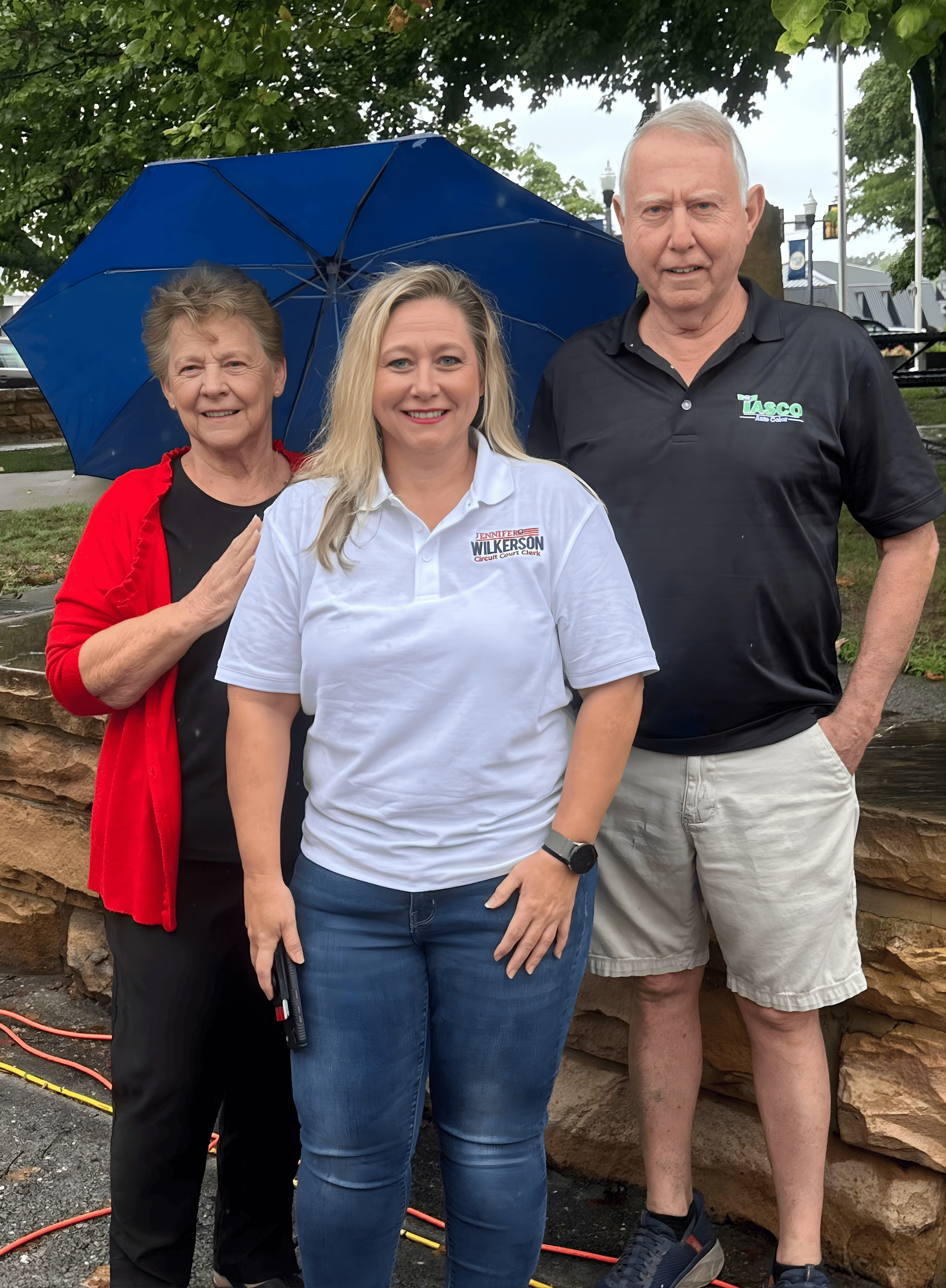 Jennifer Wilkerson standing outdoors on a cloudy day, with one holding a blue umbrella. The woman in the middle is wearing a white polo shirt with campaign logos, and the other two are dressed casually.