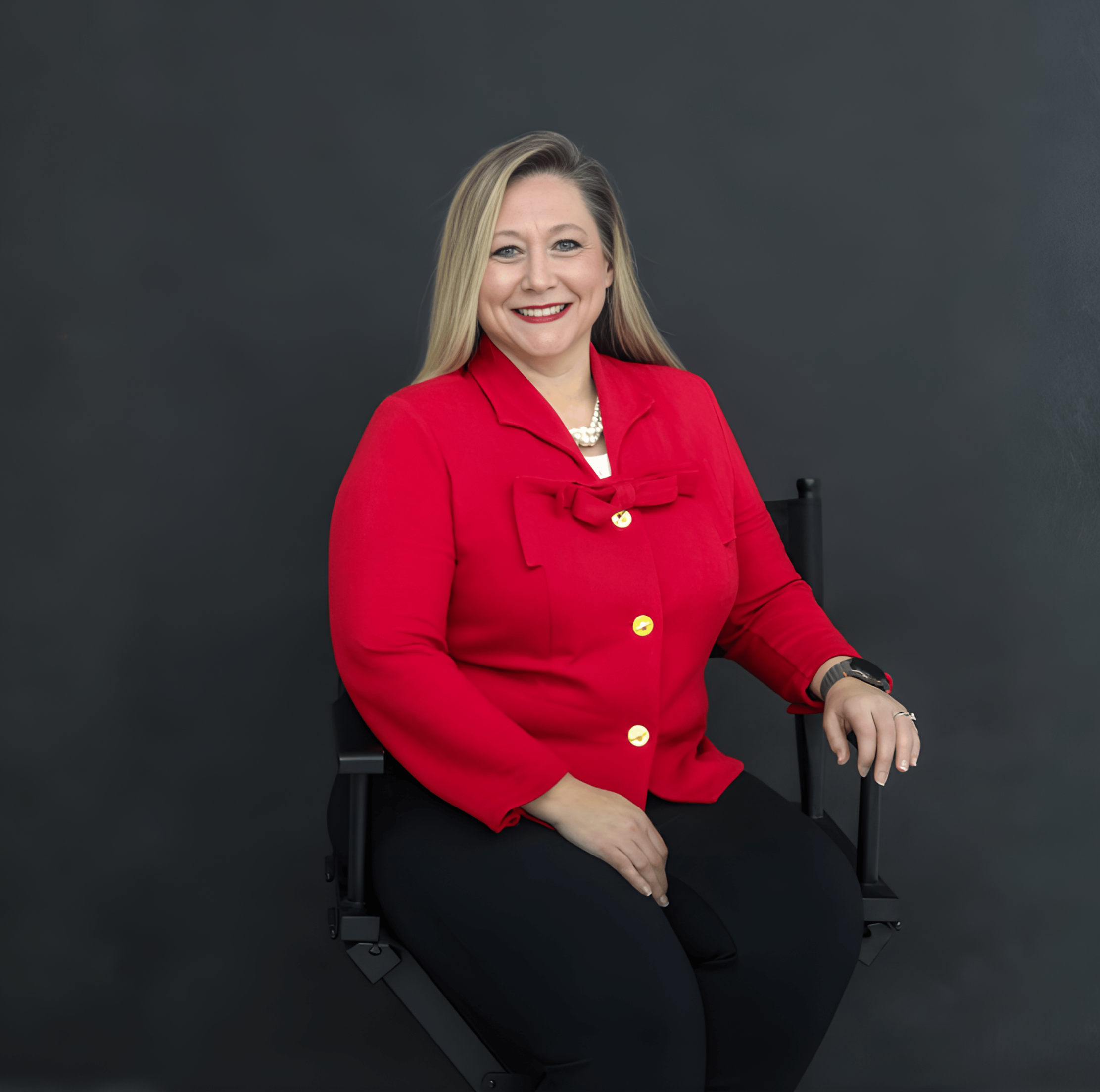 Jennifer Wilkerson with blonde hair wearing red blazer, black pants, and pearl necklace sitting on a black chair in front of a dark gray background.