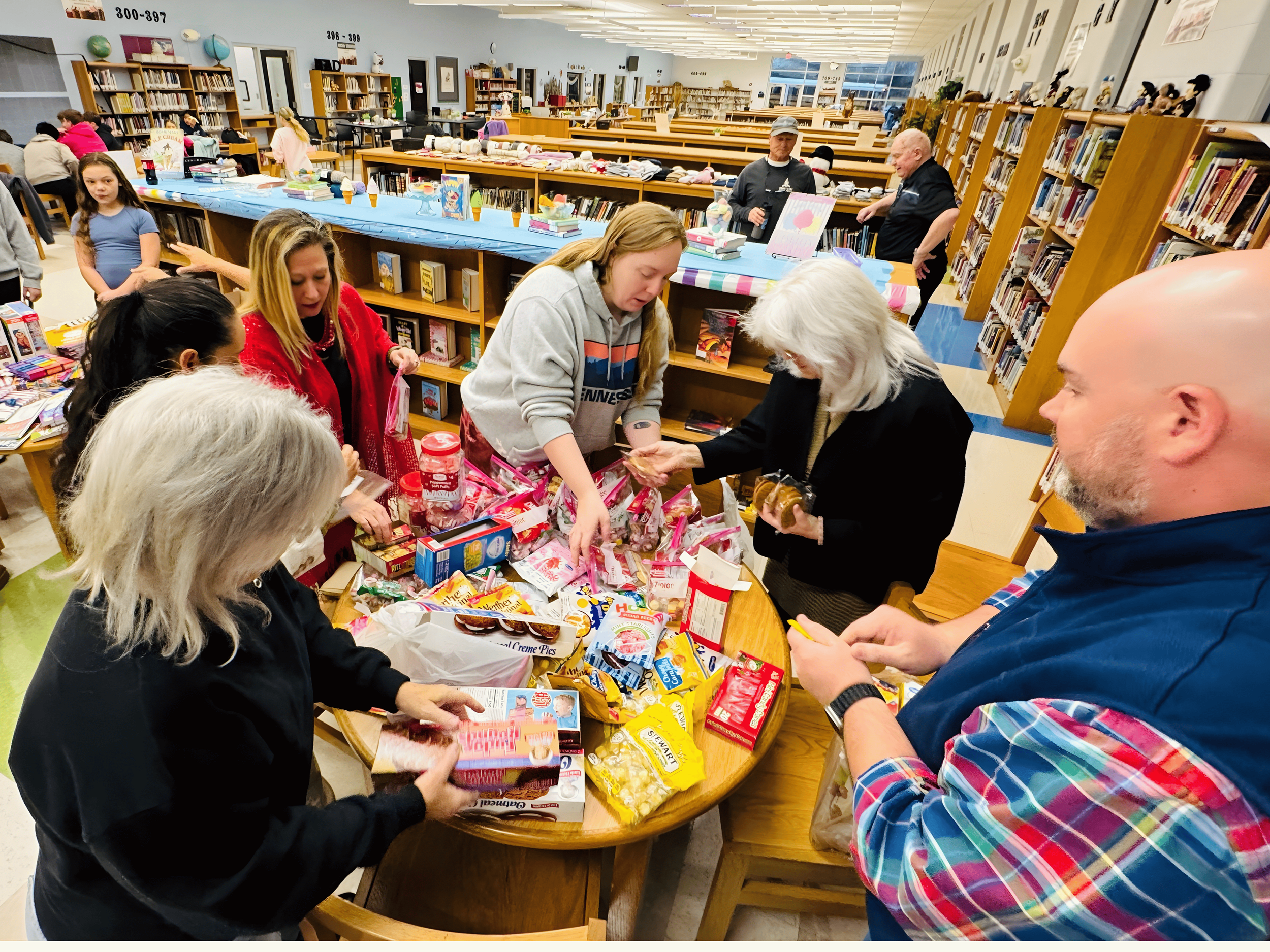 People with Jennifer Wilkerson gathered around a table in a library, exchanging and browsing various baked goods and snacks, with bookshelves and other patrons in the background.