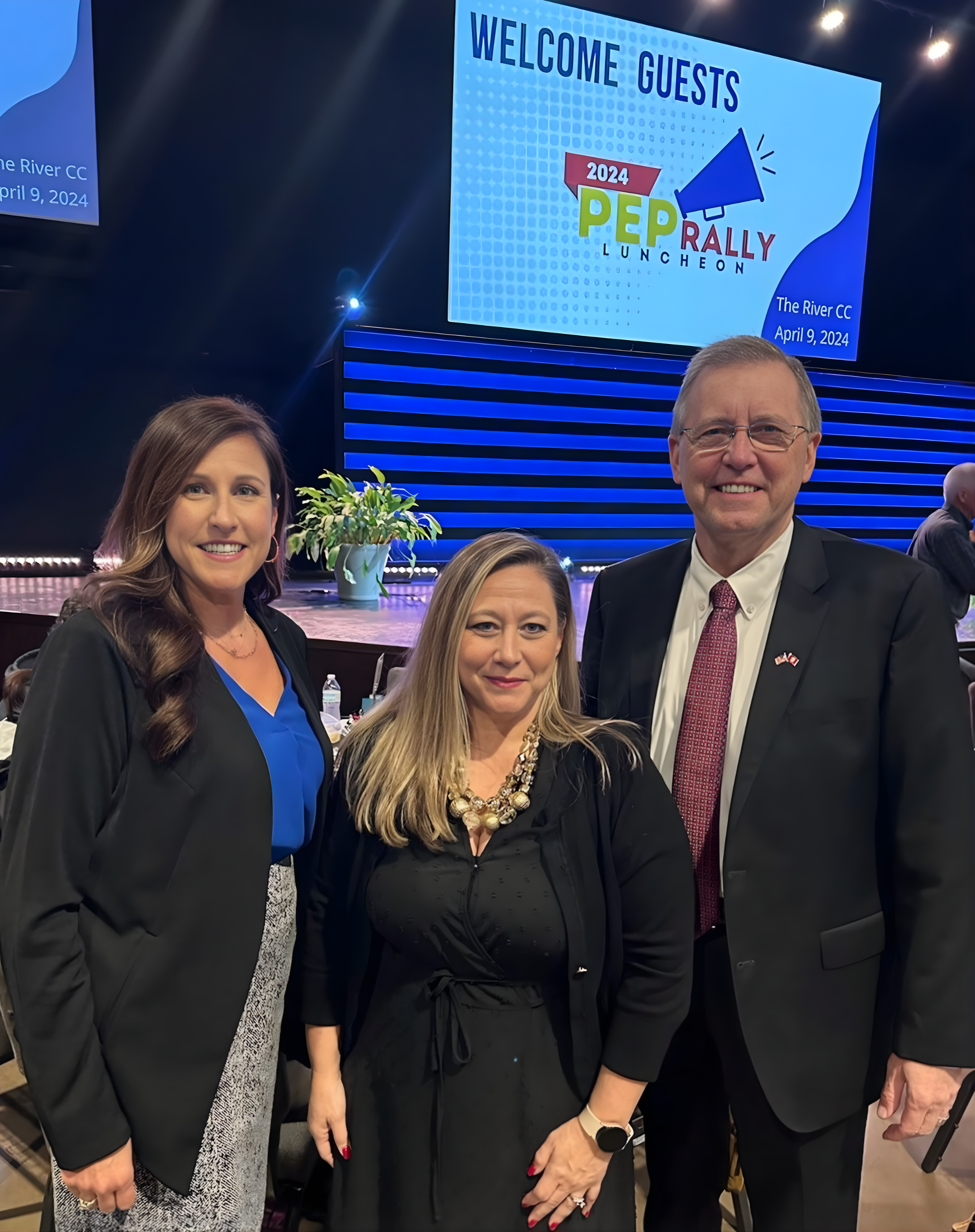 Jennifer Wilkerson posing in front of a stage with a large screen that reads 'Welcome Guests' at a rally luncheon event. The event is at The River CC on April 9, 2024. The stage has a potted plant and blue lighting.