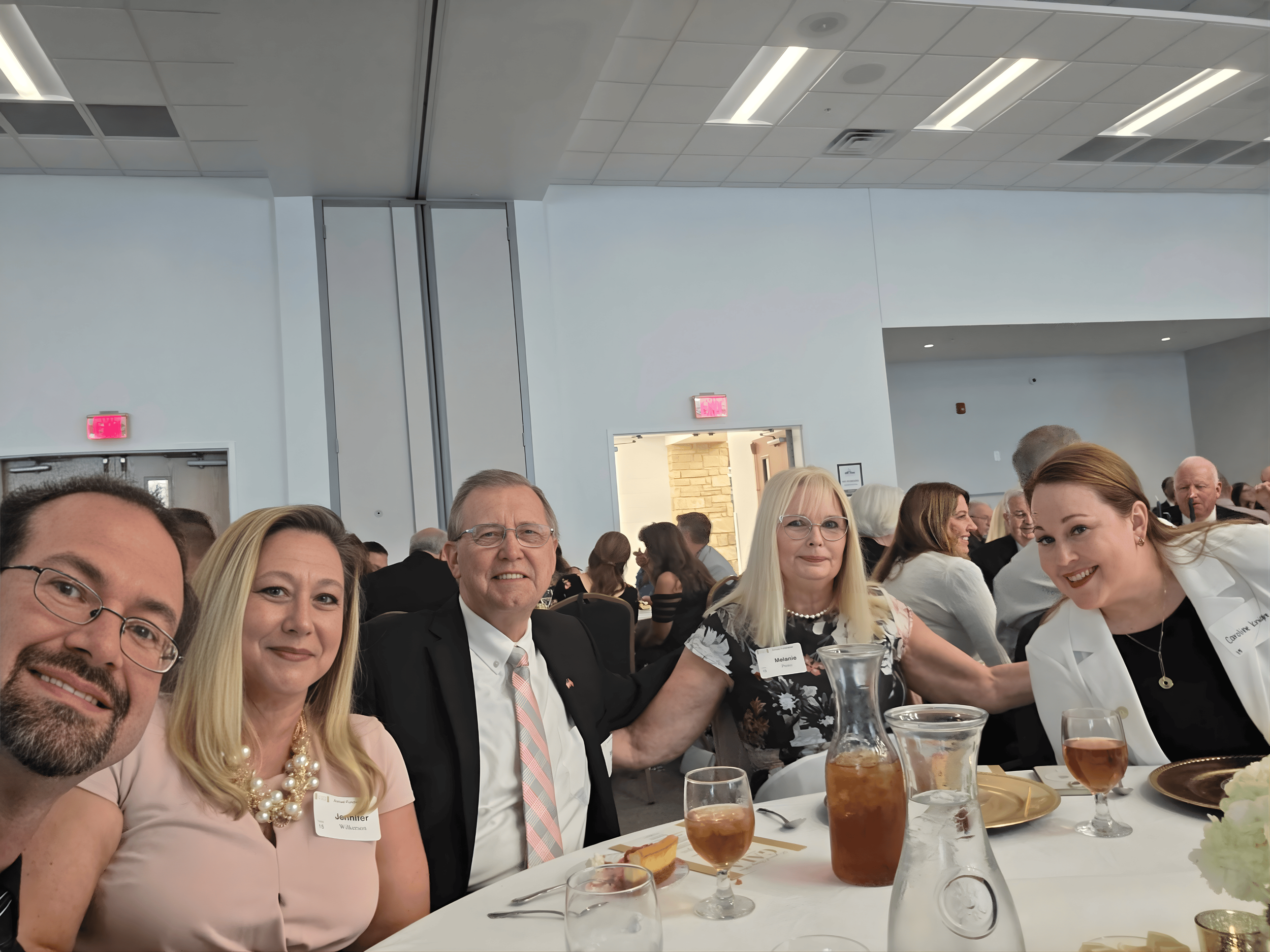 Group of five people sitting at a table during a formal event, smiling at the camera, with drinks and tableware in front of them, in a bright and modern banquet hall.