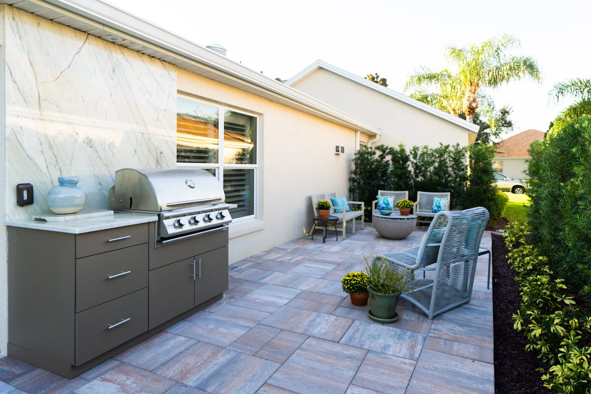Backyard patio with outdoor grill, potted flowers, wicker chairs, and garden bushes in sunny weather.