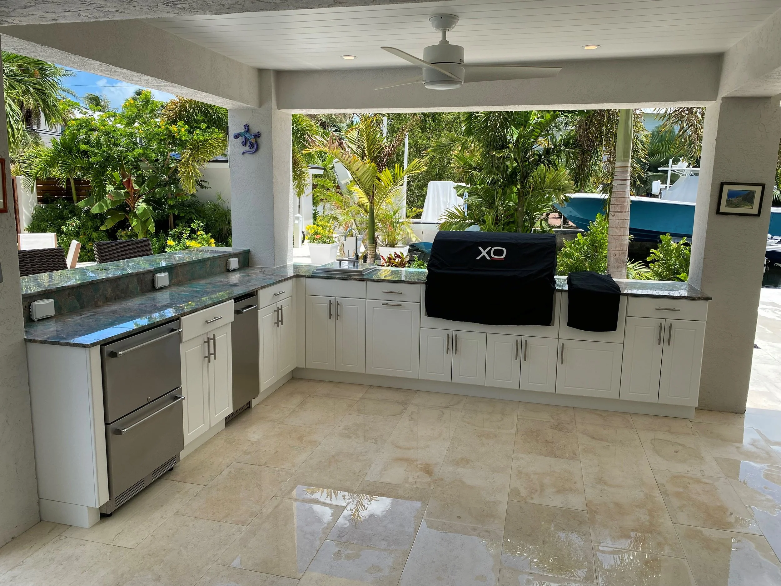 Outdoor kitchen with white cabinets, marble countertop, stainless steel appliances, and a view of lush greenery and boats outside.