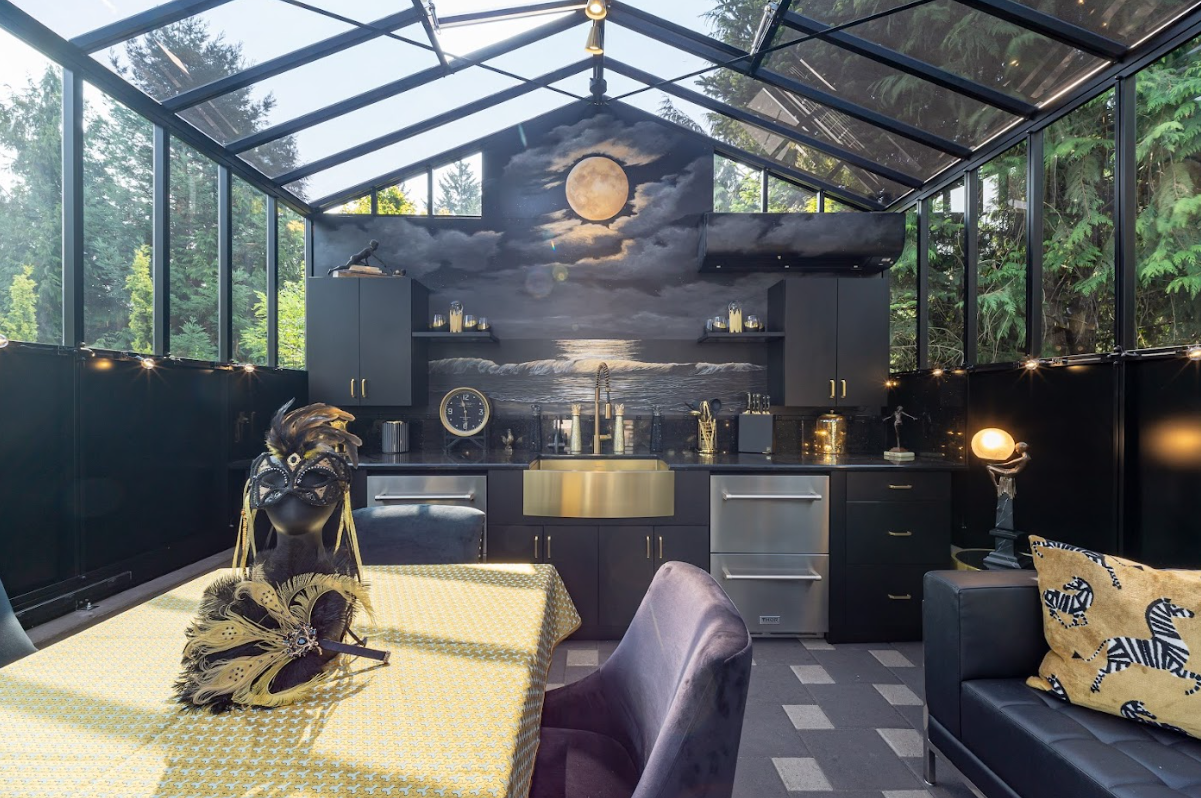 Enclosed outdoor kitchen with black cabinets, a mural of the moon over the ocean, a dining table with a decorative mask and feathers, and a black sofa with a cheetah print pillow.