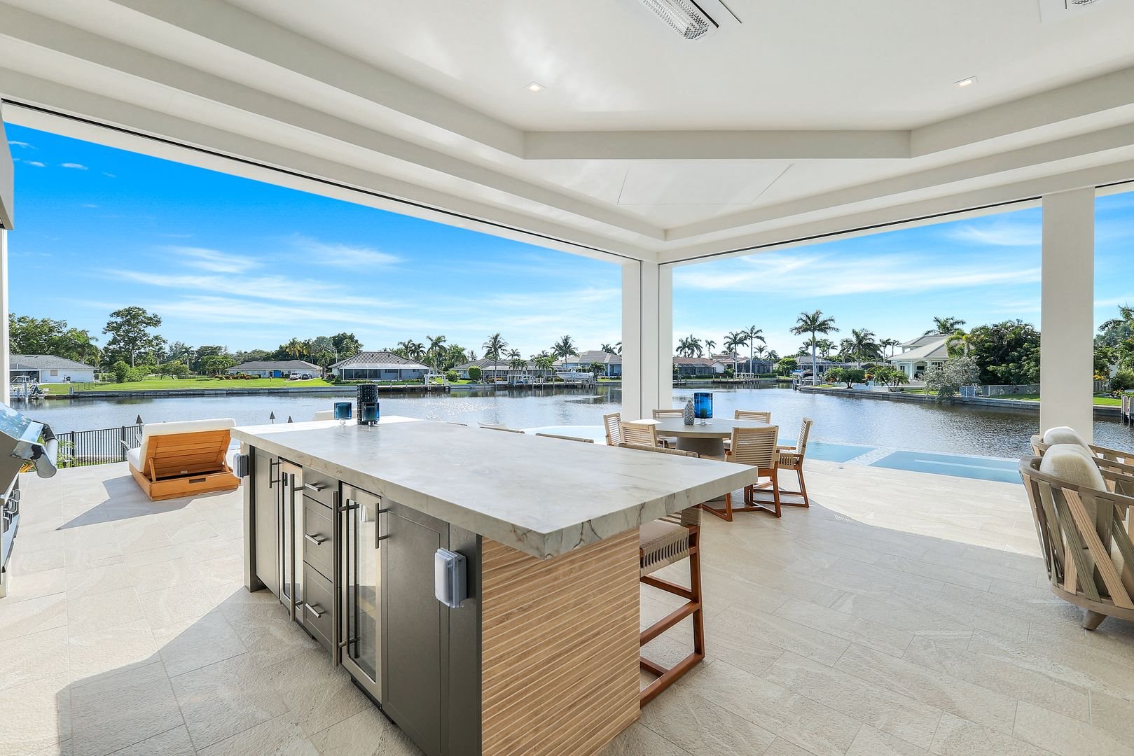 Open patio area overlooking a waterway with houses and palm trees in the background, featuring a kitchen island with a marble countertop and wooden bar stools, a round dining table with chairs, and lounge chairs nearby.