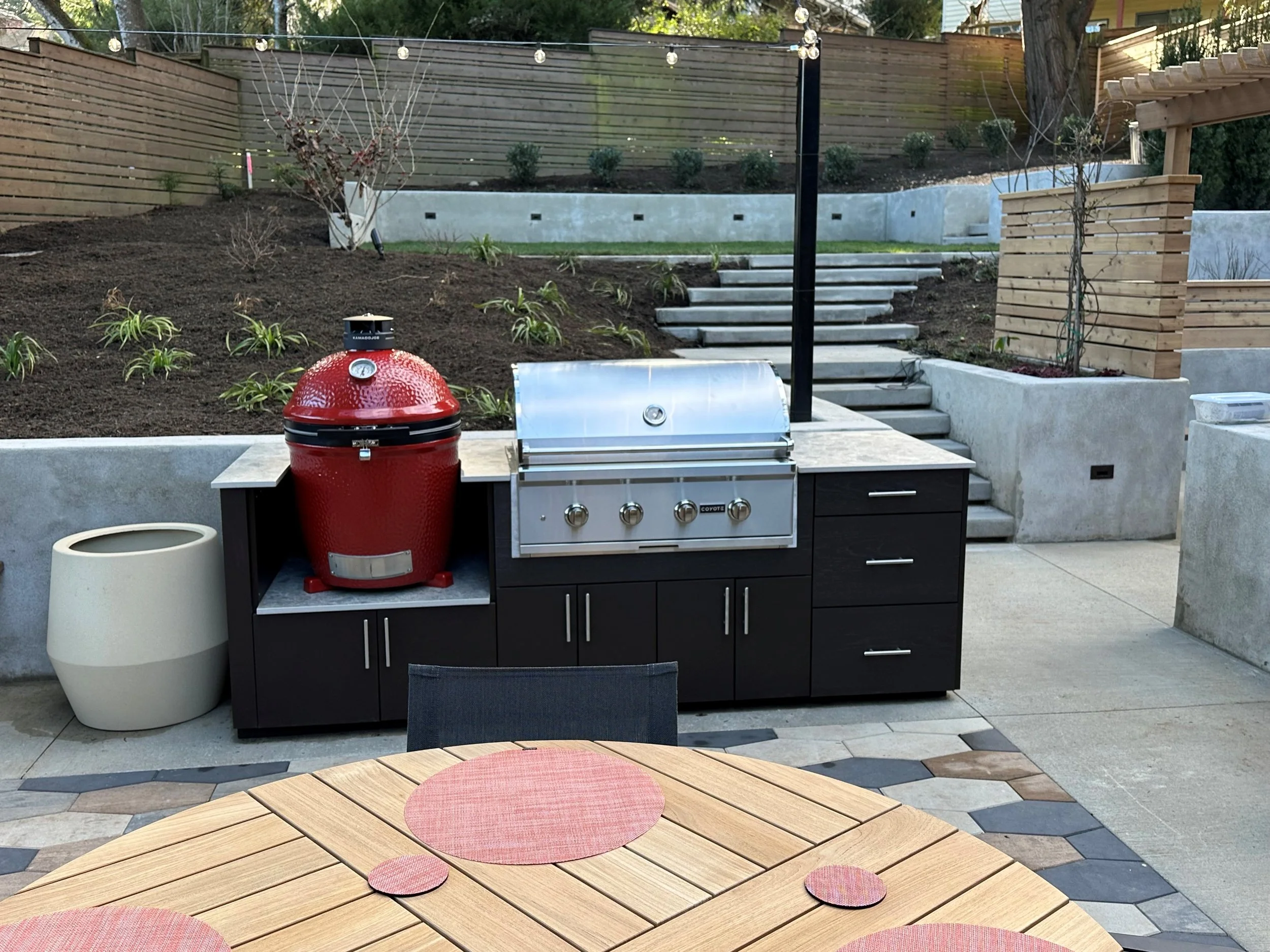 Outdoor backyard kitchen area with a red Kamado-style grill, stainless steel gas grill, black cabinetry, and surrounding planters and landscaping.