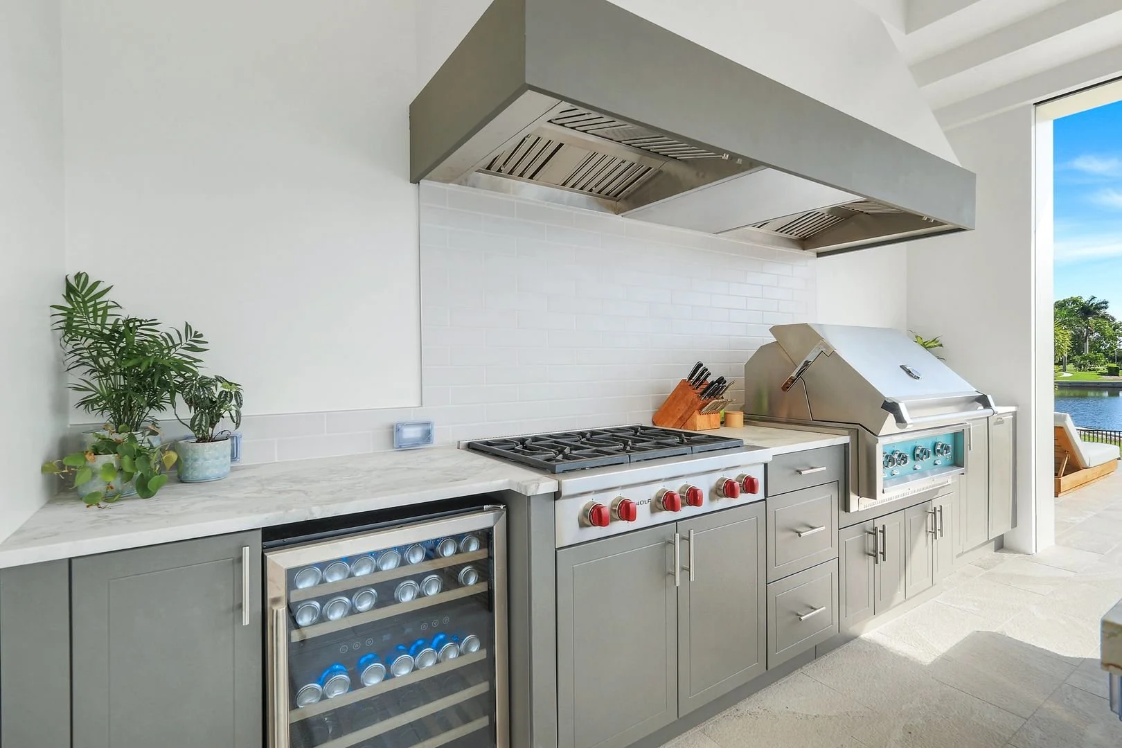 Outdoor kitchen with gray cabinetry, a white marble countertop, a gas stove with red knobs, a built-in wine cooler, and a stainless steel grill. There are potted plants on the countertop and a large window showing a lakeside outdoor area.