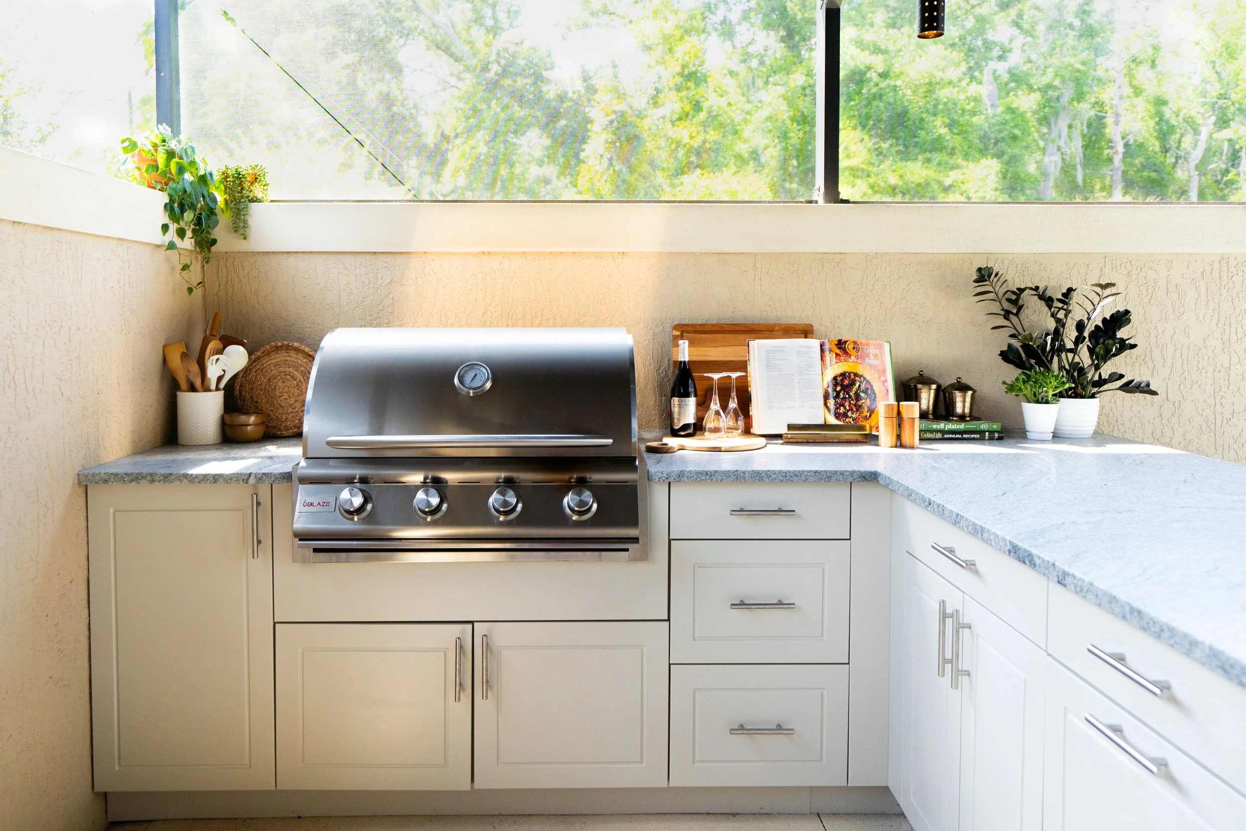 Outdoor kitchen with a stainless steel gas grill, potted plants, cookbooks, and wine bottles on a gray granite countertop, with a window showing trees outside.