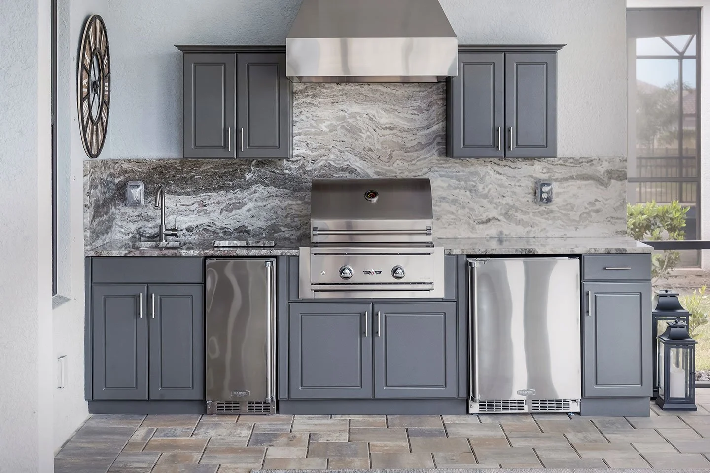 Modern outdoor kitchen with gray cabinets, marble backsplash, stainless steel grill, mini fridge, and large window