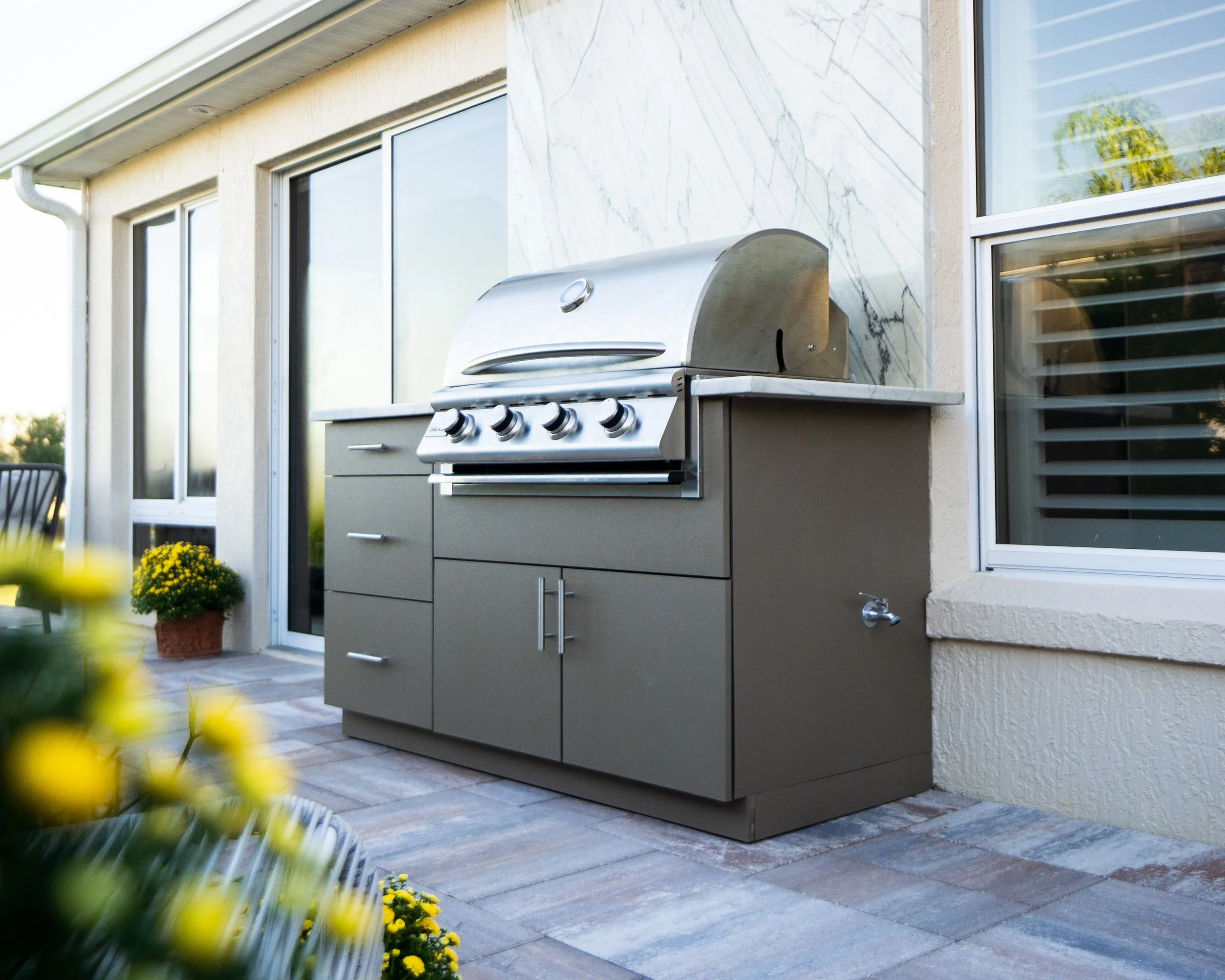 Outdoor stainless steel gas grill on a patio with potted yellow flowers and sliding glass doors.