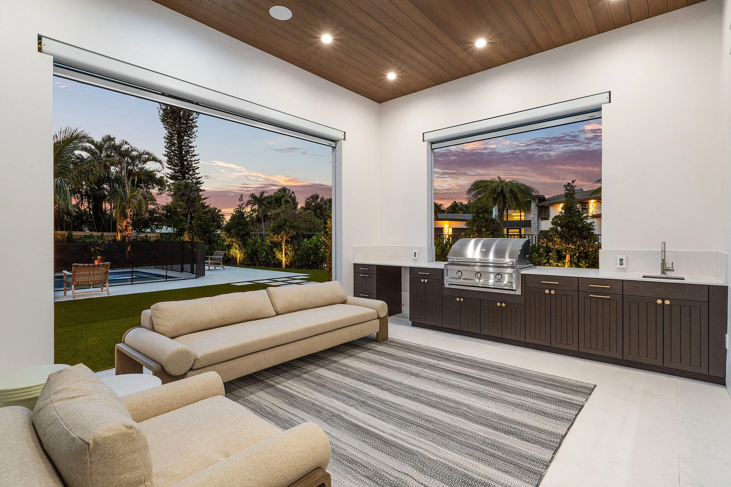 View of a modern indoor living space with large windows showing an outdoor backyard with a pool, trees, and sunset sky. Inside, there are beige sofas, a striped rug, a built-in grill, cabinetry, and a countertop.