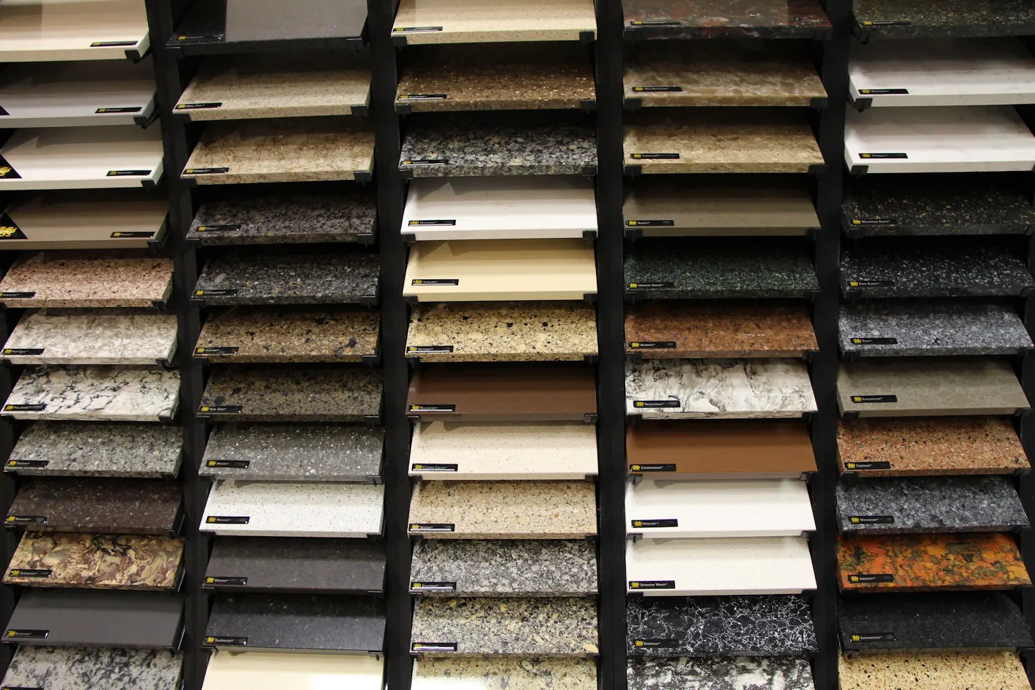 Display of various countertop samples in different colors and patterns arranged on black shelving units.