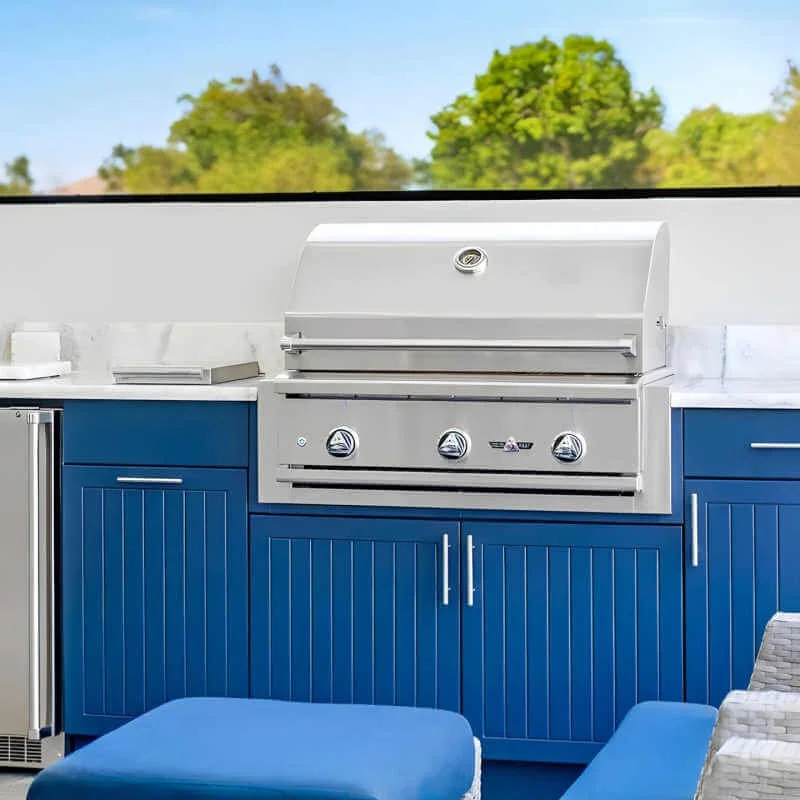 A modern outdoor kitchen with a stainless steel grill set into blue cabinetry, situated beneath a window showing green trees outside.