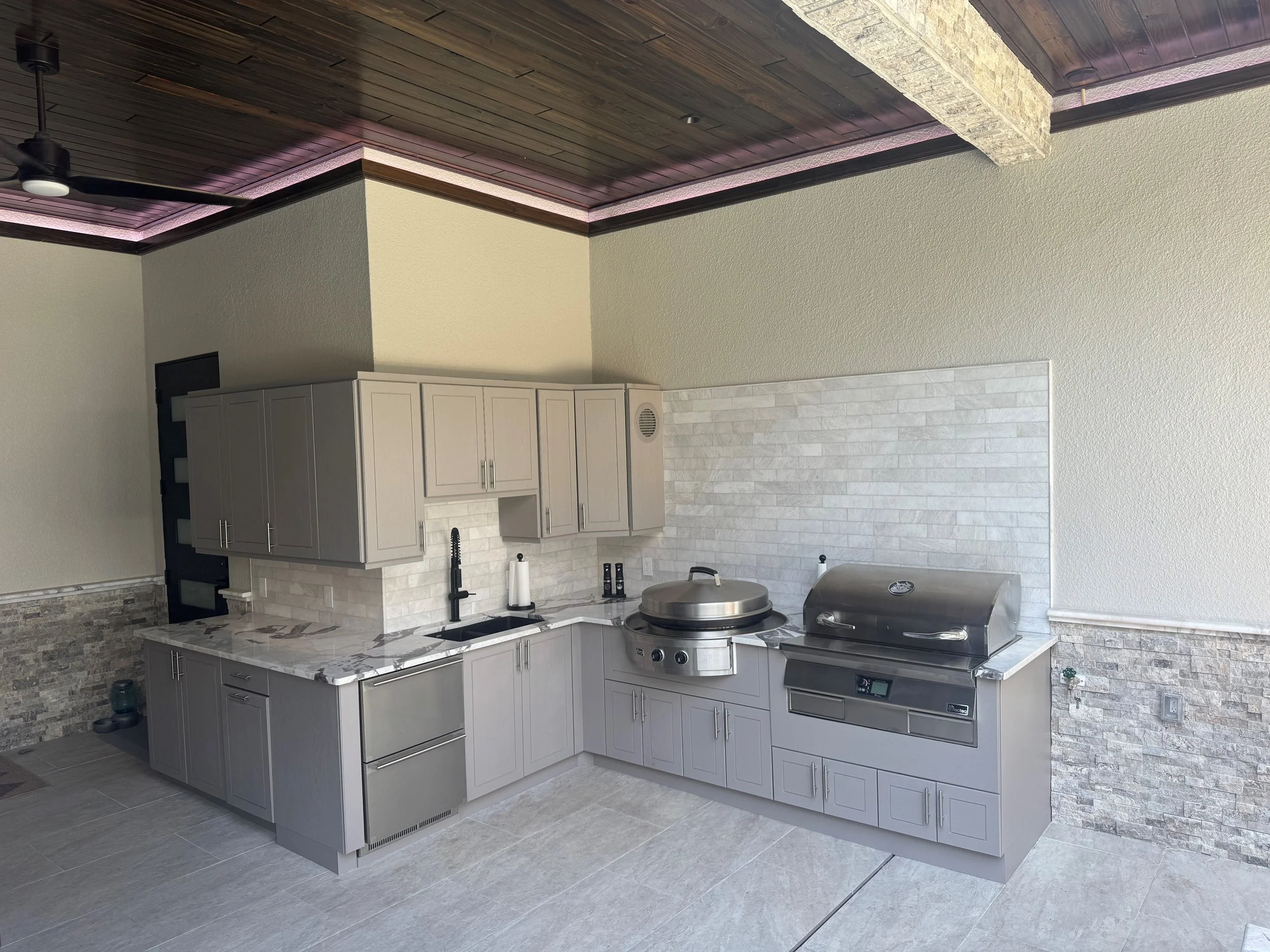 Outdoor kitchen with gray cabinets, white marble countertops, and stainless steel grilling appliances, including a large grill and a rounded griddle, against a white brick backsplash and beige stone accents.