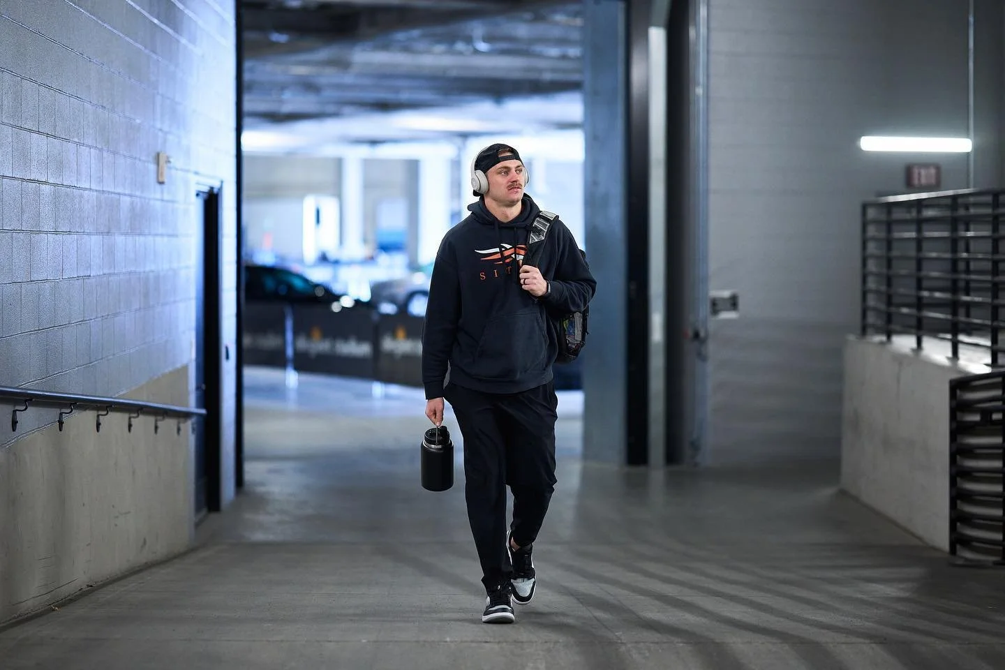 A young man wearing a black hoodie with a logo, black pants, sneakers, and a backwards cap, walking through an underpass while carrying a water bottle in one hand and headphones on his head. He is also carrying a backpack.