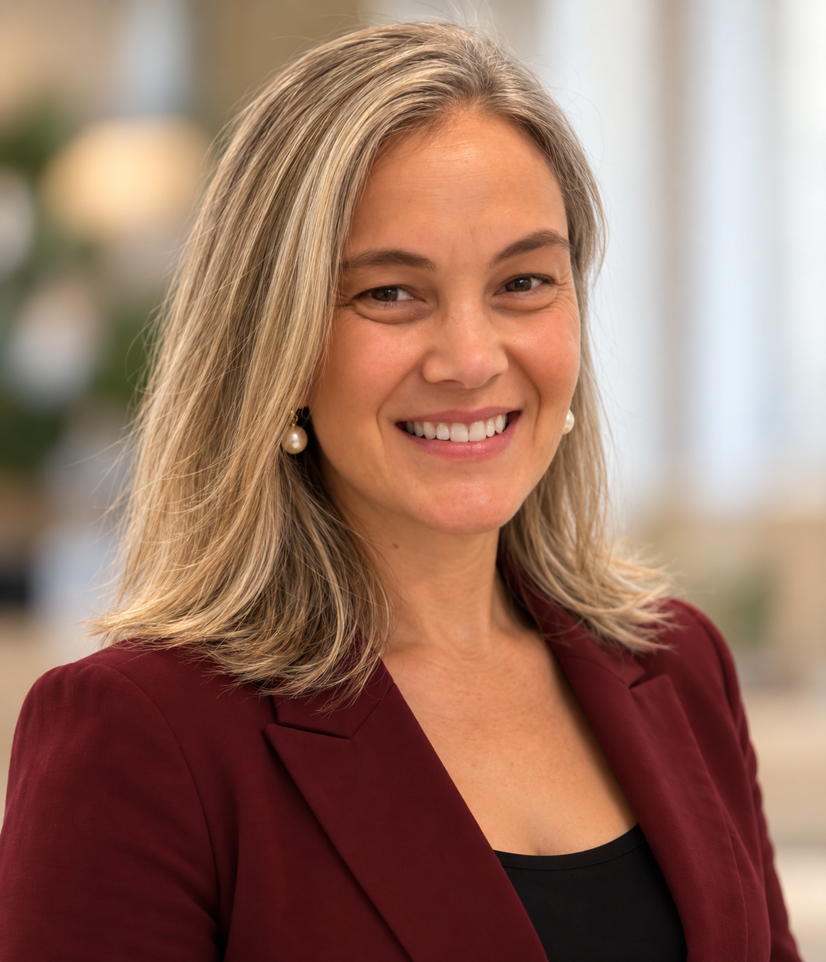 A woman with shoulder-length blonde hair, wearing pearl earrings and a maroon blazer, smiling in an indoor setting.