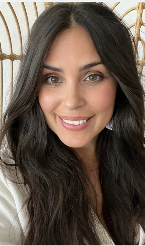 A woman with long dark wavy hair, smiling, wearing a black sleeveless button-up dress, standing in front of a white curtain.