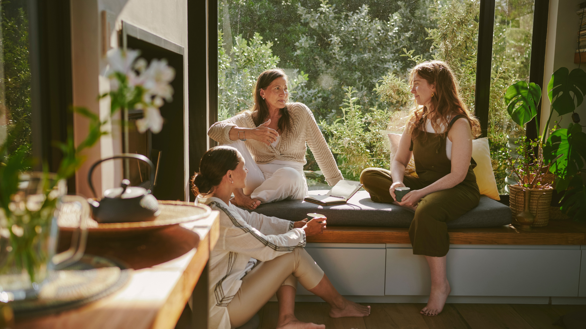 Four women having a conversation in a sunlit room with large windows and plants, sitting on a window seat with a book open.