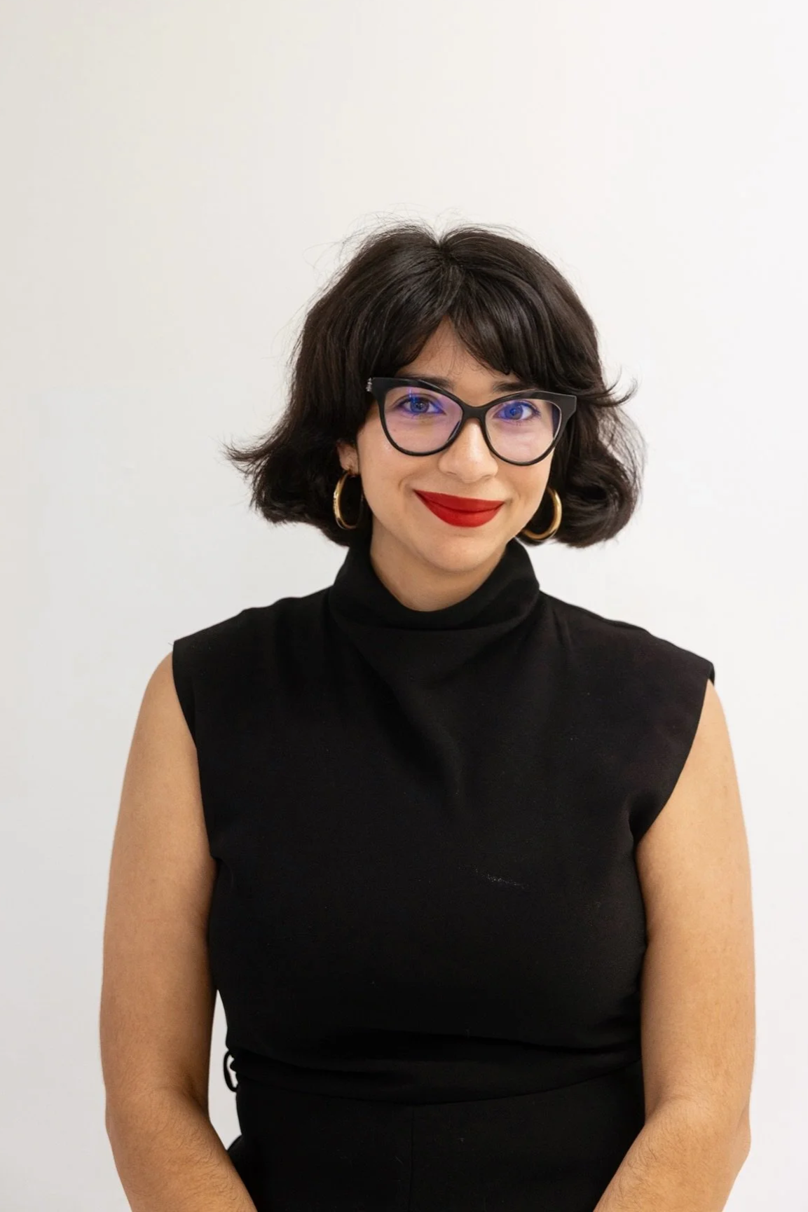 A woman with short dark hair, wearing glasses, gold hoop earrings, red lipstick, and a brown turtleneck sweater, seated indoors near a window with a Christmas tree and gold ornaments visible outside. There is a floral arrangement with orange and white flowers on a wooden cabinet behind her.
