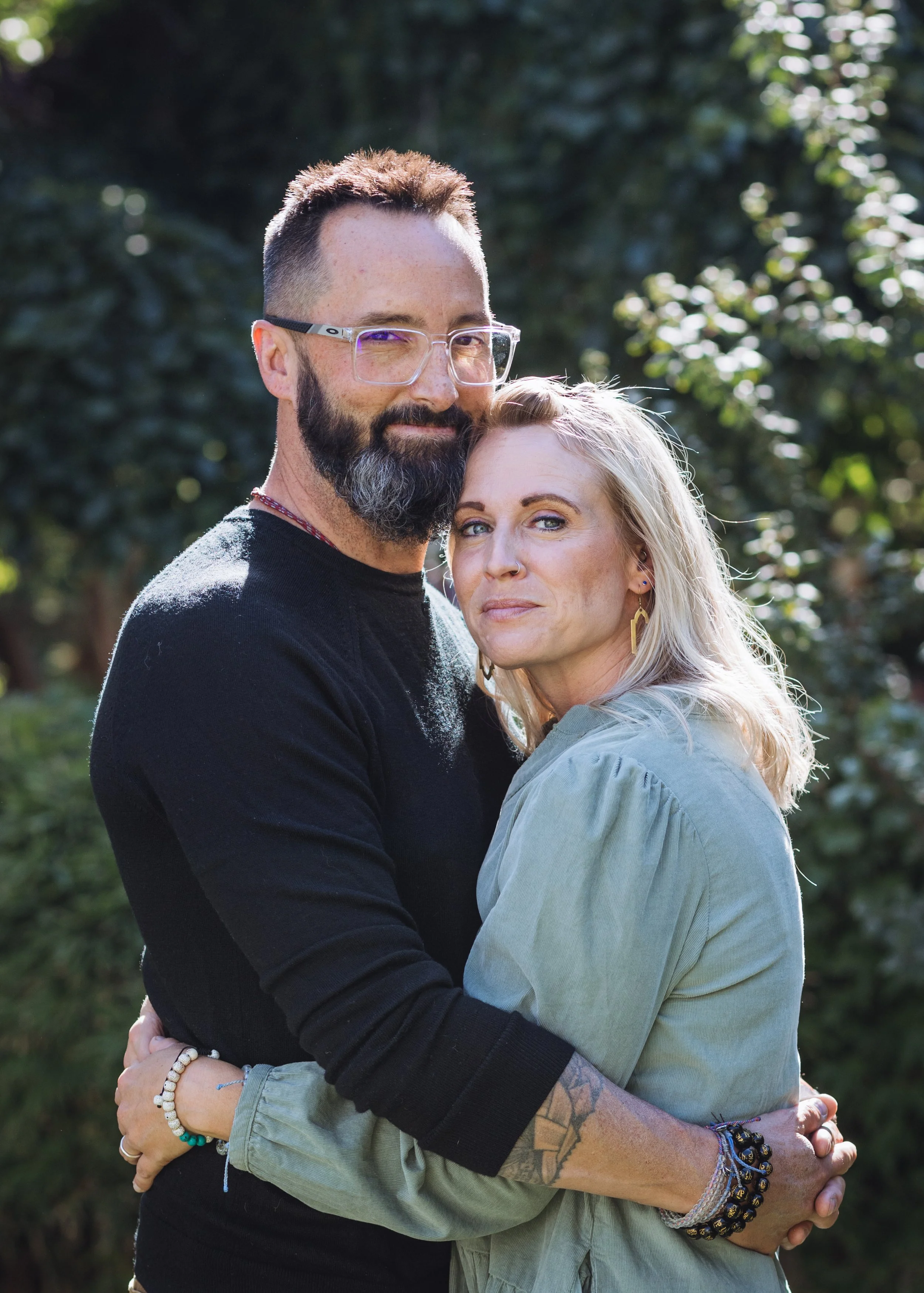 A man and woman embracing outdoors, with trees and sunlight in the background.