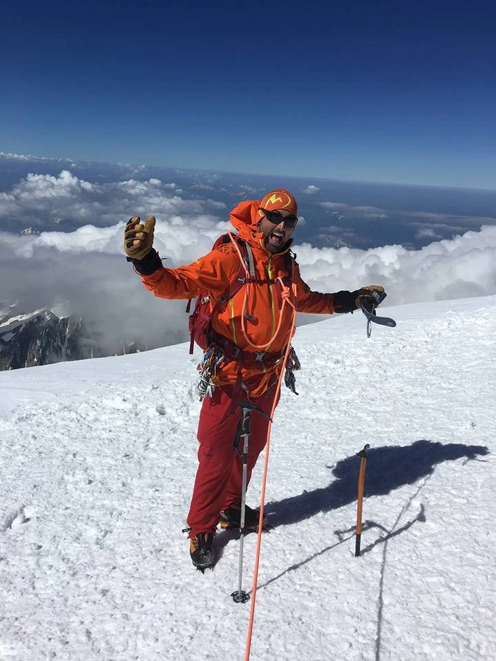 A person standing on a snow-covered mountain peak, dressed in orange and red mountaineering gear, with climbing tools in hand, smiling with arms raised, against a backdrop of clouds and blue sky.