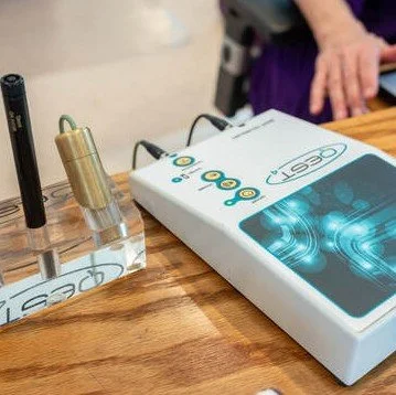 Close-up of a scientific testing device and a box on a wooden table, with a person's hand in the background.