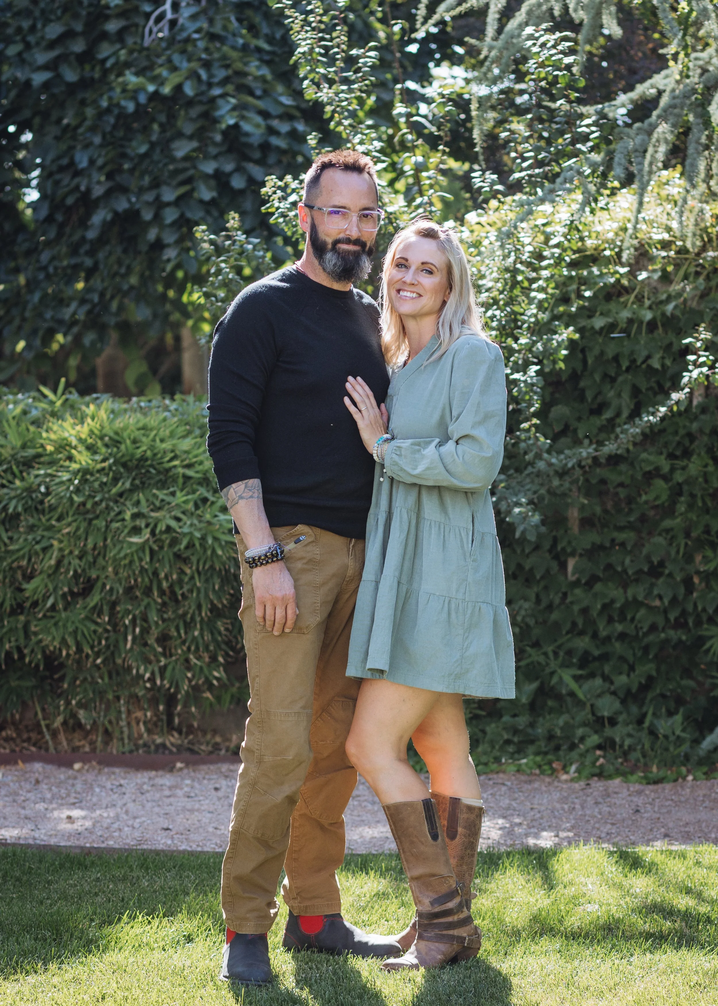 A couple stands outdoors in sunlight, surrounded by lush greenery, smiling at the camera. The man has a beard, glasses, and tattoos, and is wearing a black sweater and khaki pants. The woman has blonde hair, and is wearing a light green dress and brown boots. They are close together, with the woman's hand on the man's chest.