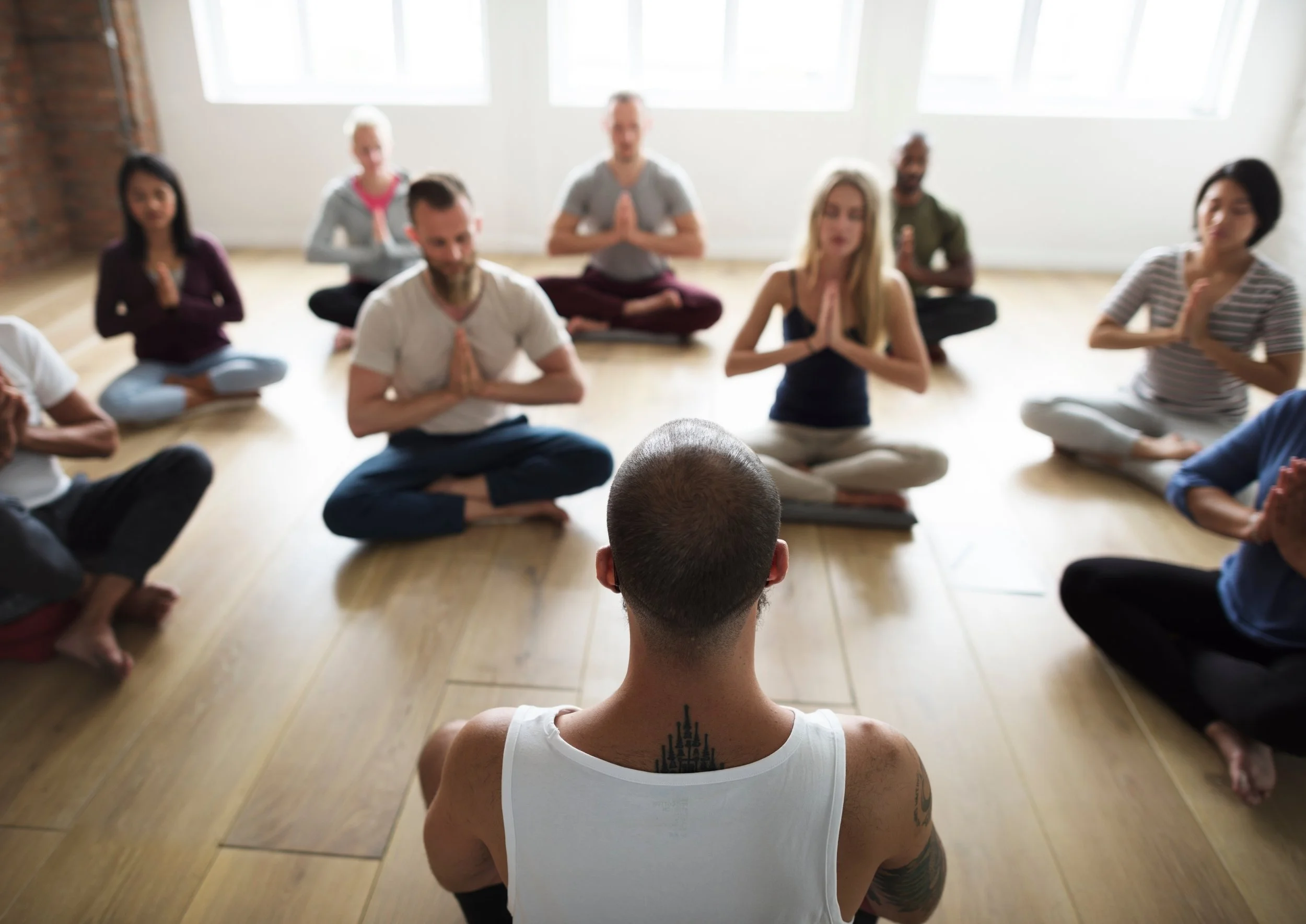 A group of people practicing meditation in a yoga class, sitting on mats with their legs crossed and hands in a prayer position, in front of a teacher.