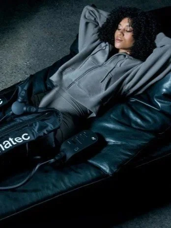 Woman lying on a leather couch, wearing a gray hoodie and black pants, with her eyes closed, surrounded by black leather items and electronics.
