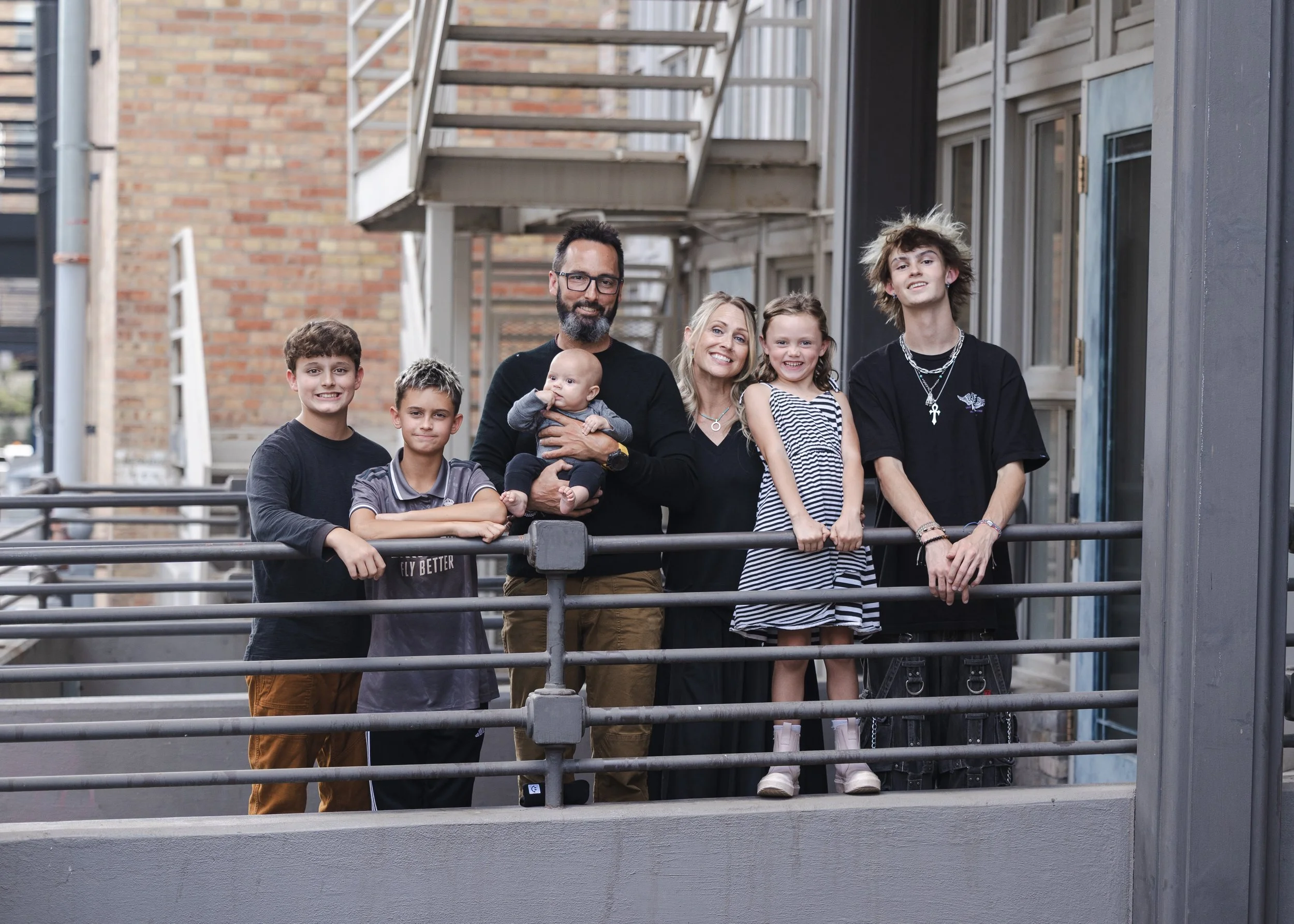A family of seven posing on a city outdoor balcony, with brick buildings behind them. The group includes three boys, two girls, a man holding a baby, and a woman. They are all smiling and casually dressed.