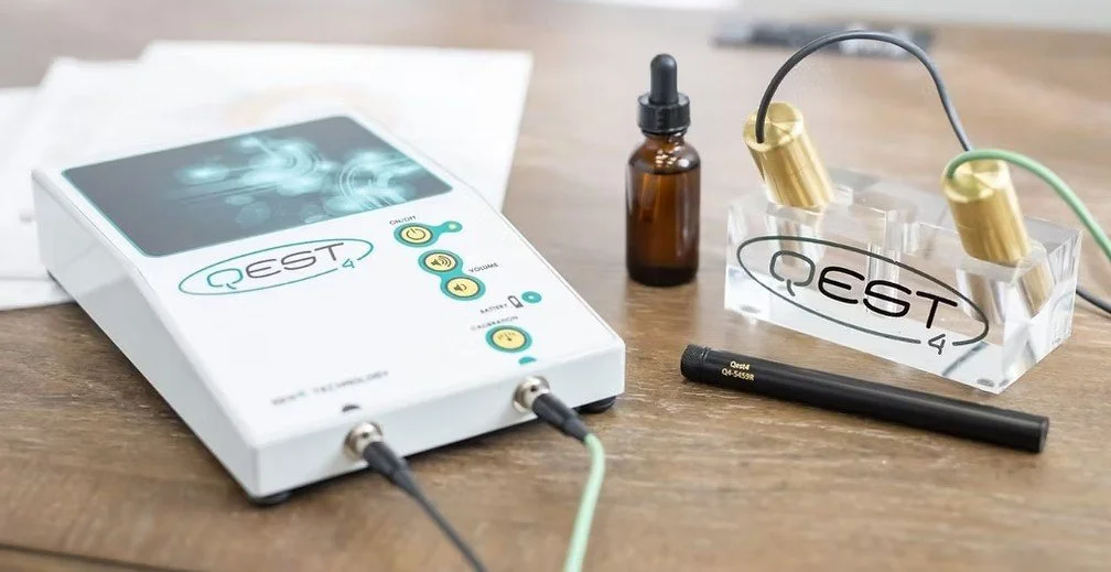 Medical device with a black cable attached, a small brown bottle with a dropper, a clear plastic container with two gold electrodes, and a black pen-like object on a wooden surface.