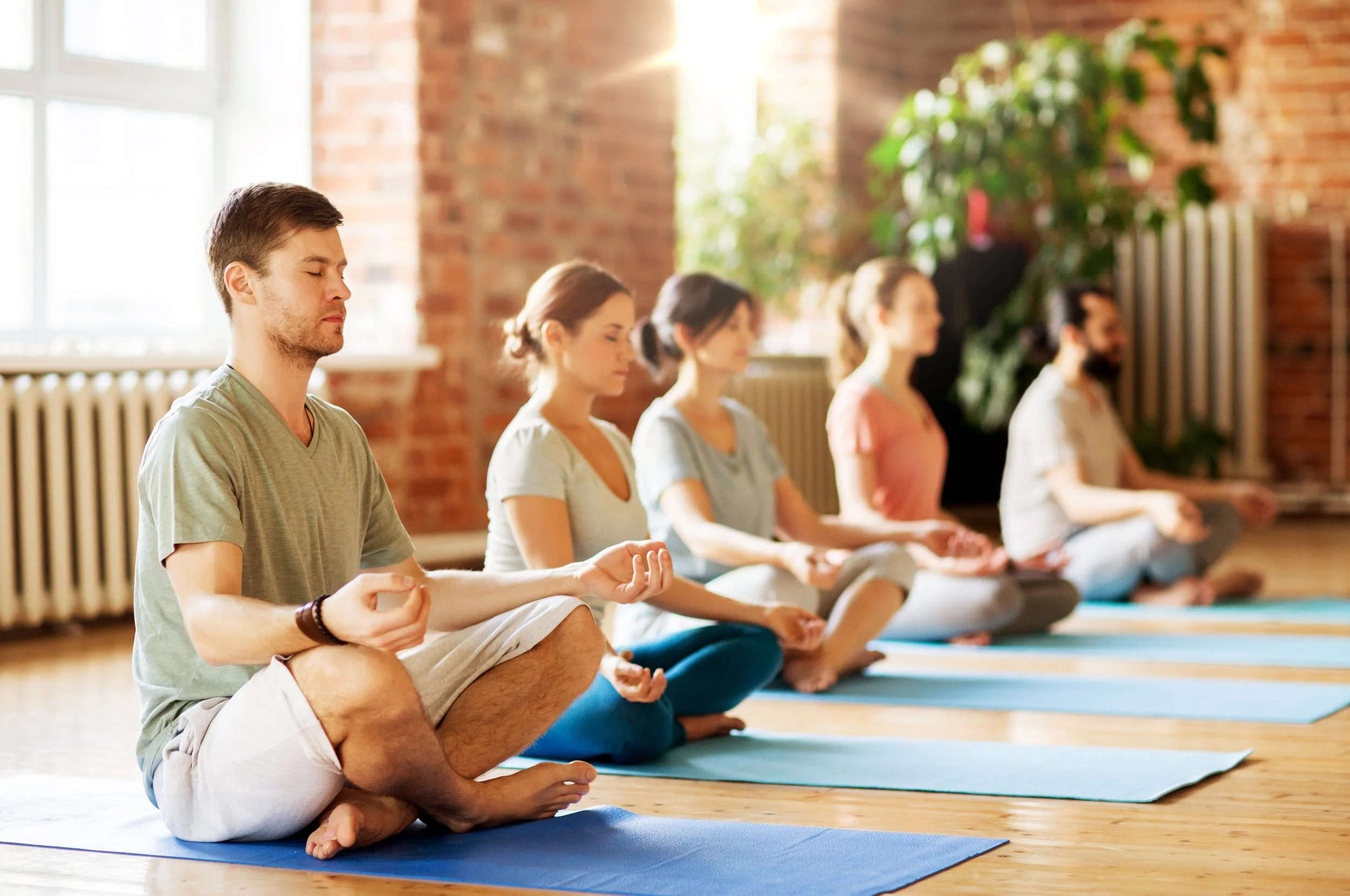 Group of people practicing yoga in a bright studio with brick walls, sitting cross-legged in meditation, focusing.