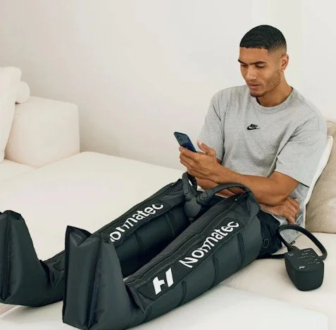 A young man sitting on a bed using his smartphone, with exercise equipment and a small black speaker nearby.