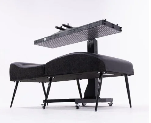 A modern massage table with a black cushioned surface and an overhead mechanical device, possibly for therapeutic purposes.