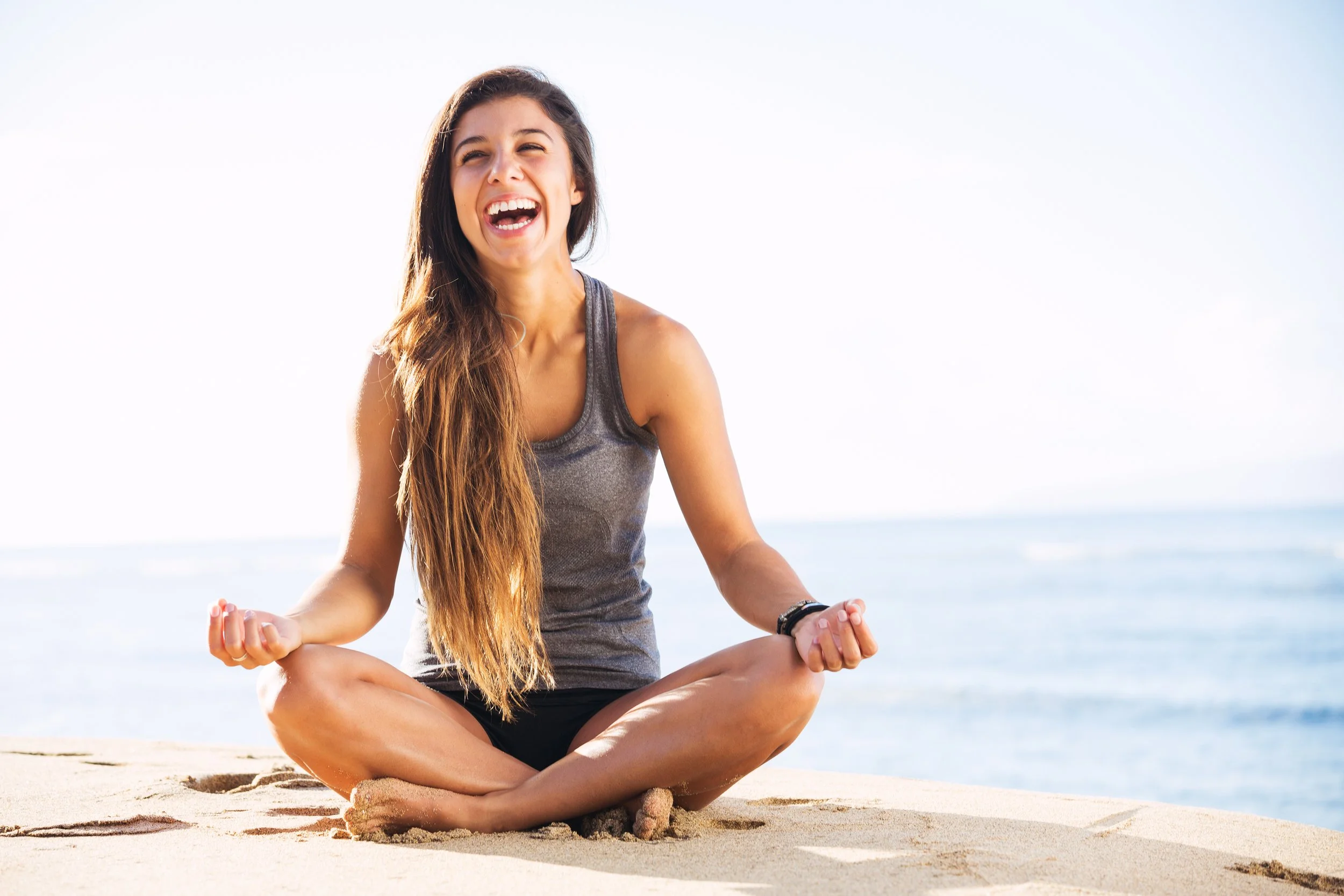 Young woman with long brown hair sitting cross-legged on the sand by the ocean, smiling and meditating.