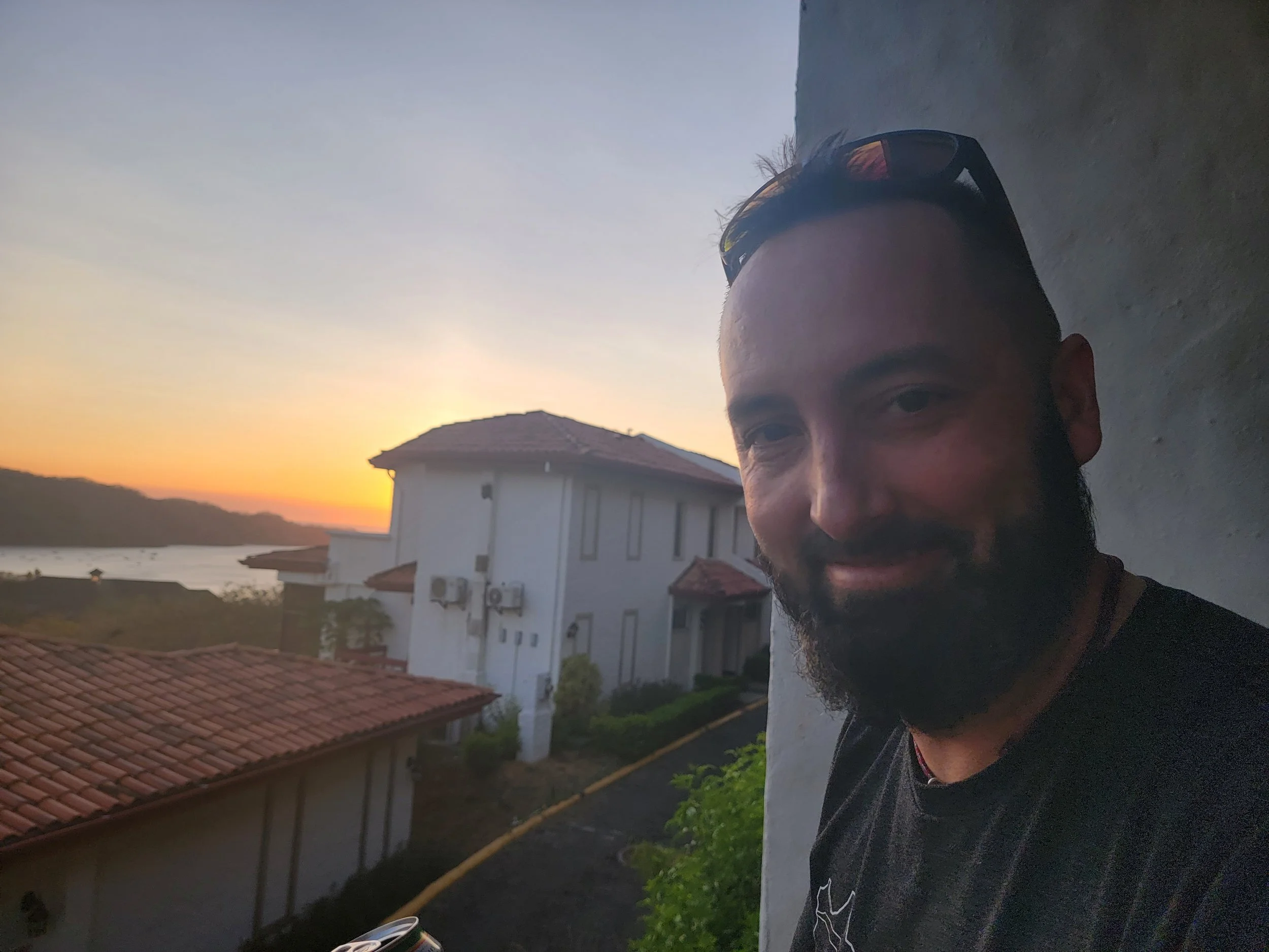 A smiling man with sunglasses resting on his head taking a selfie outdoors during sunset. Behind him, there are residential buildings with tiled roofs, greenery, a river, and hills in the distance.