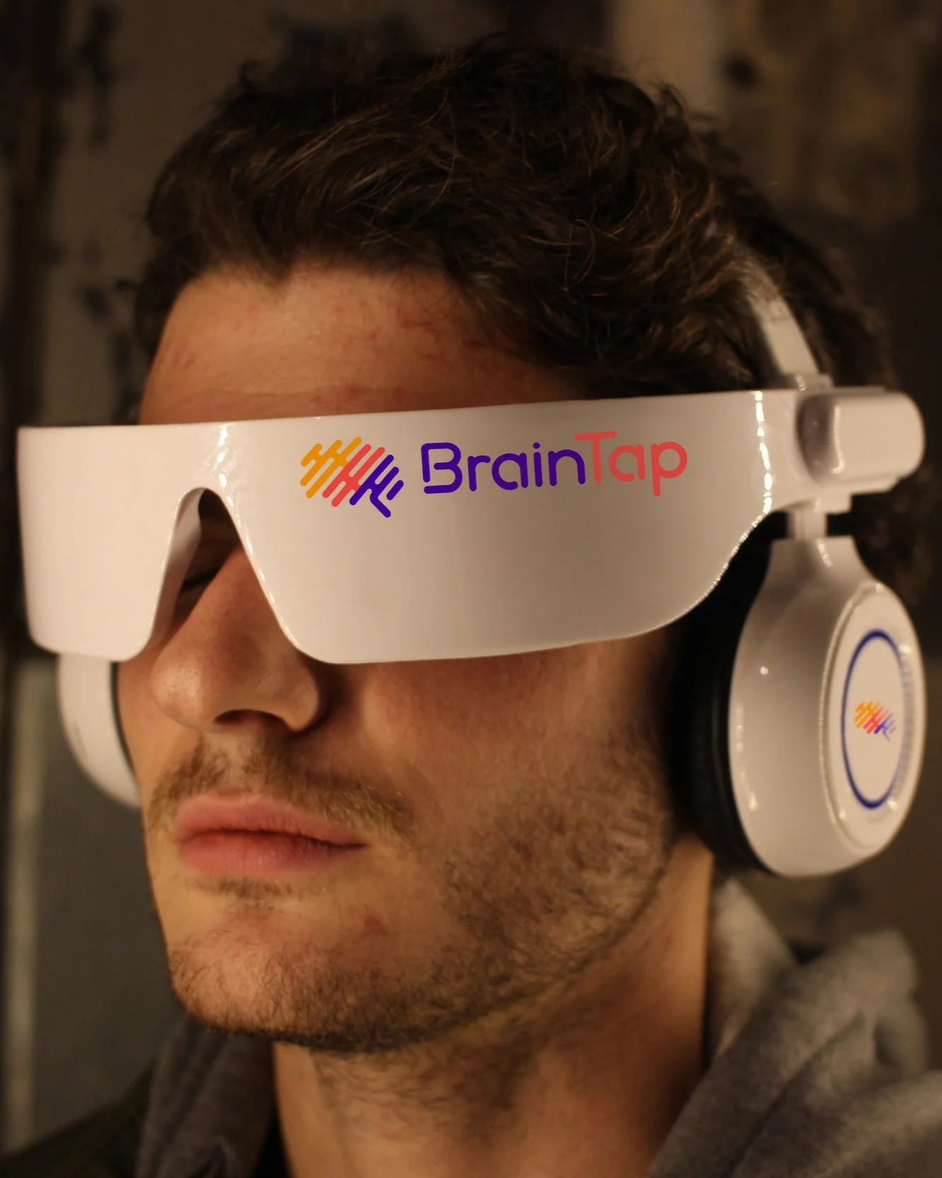 A young man wearing white VR goggles with the logo 'BrainTap' on the front, also wearing headphones, and a gray hoodie.