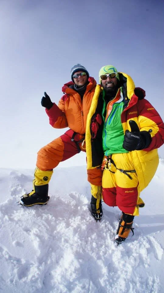 Two men standing on snow in mountaineering gear, both giving thumbs up, dressed in colorful insulated jackets and pants, with snow boots, sunglasses, and beanies.