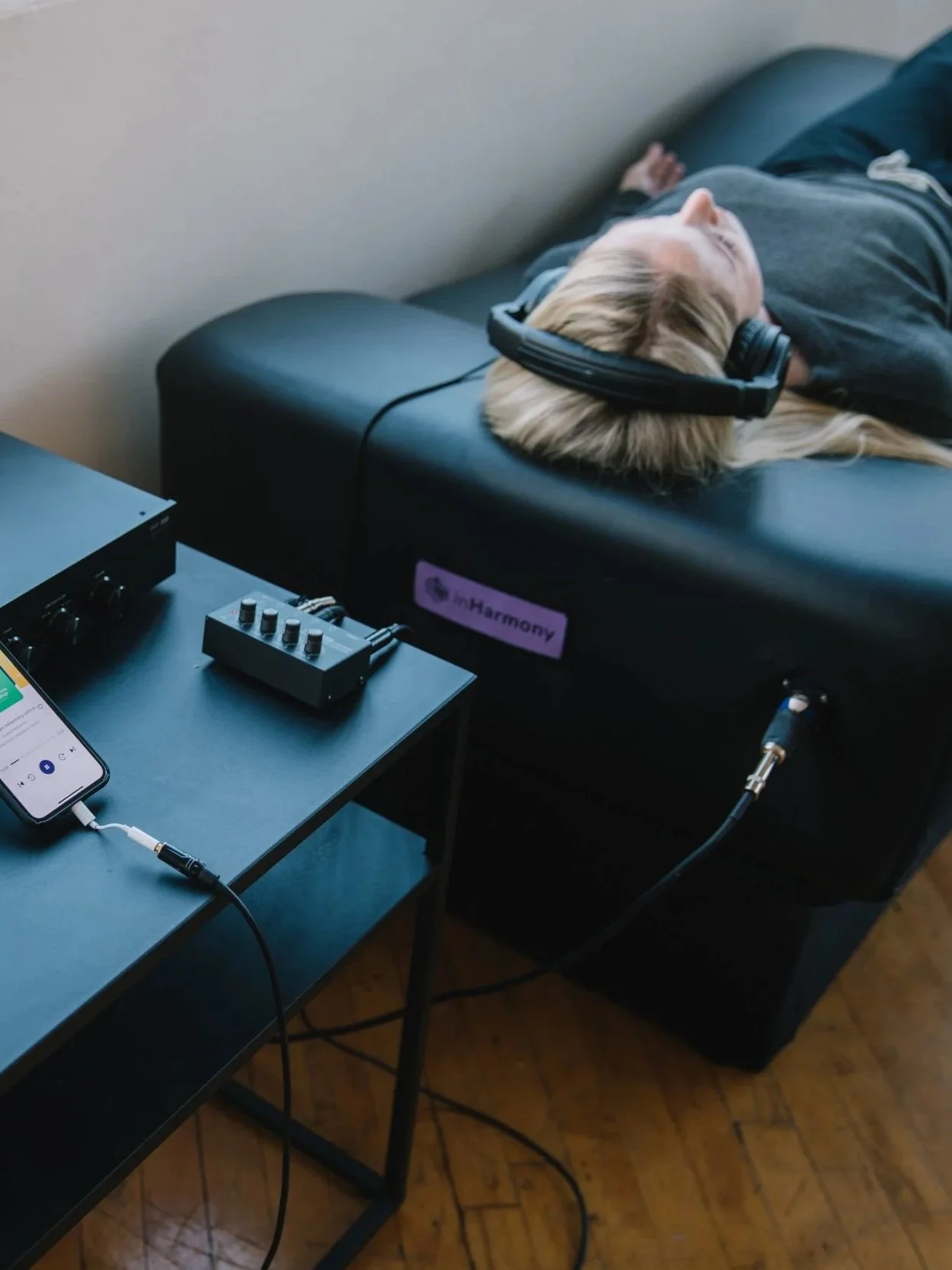 A person with long blonde hair lying on a black bed with headphones on, connected to audio equipment on a table. The person appears to be listening to music or audio using headphones.
