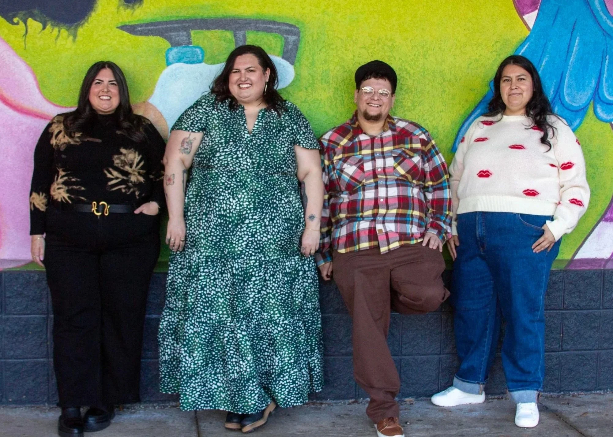 Four people standing in front of a colorful painted wall, smiling at the camera. The wall features green, pink, and blue colors with abstract designs.