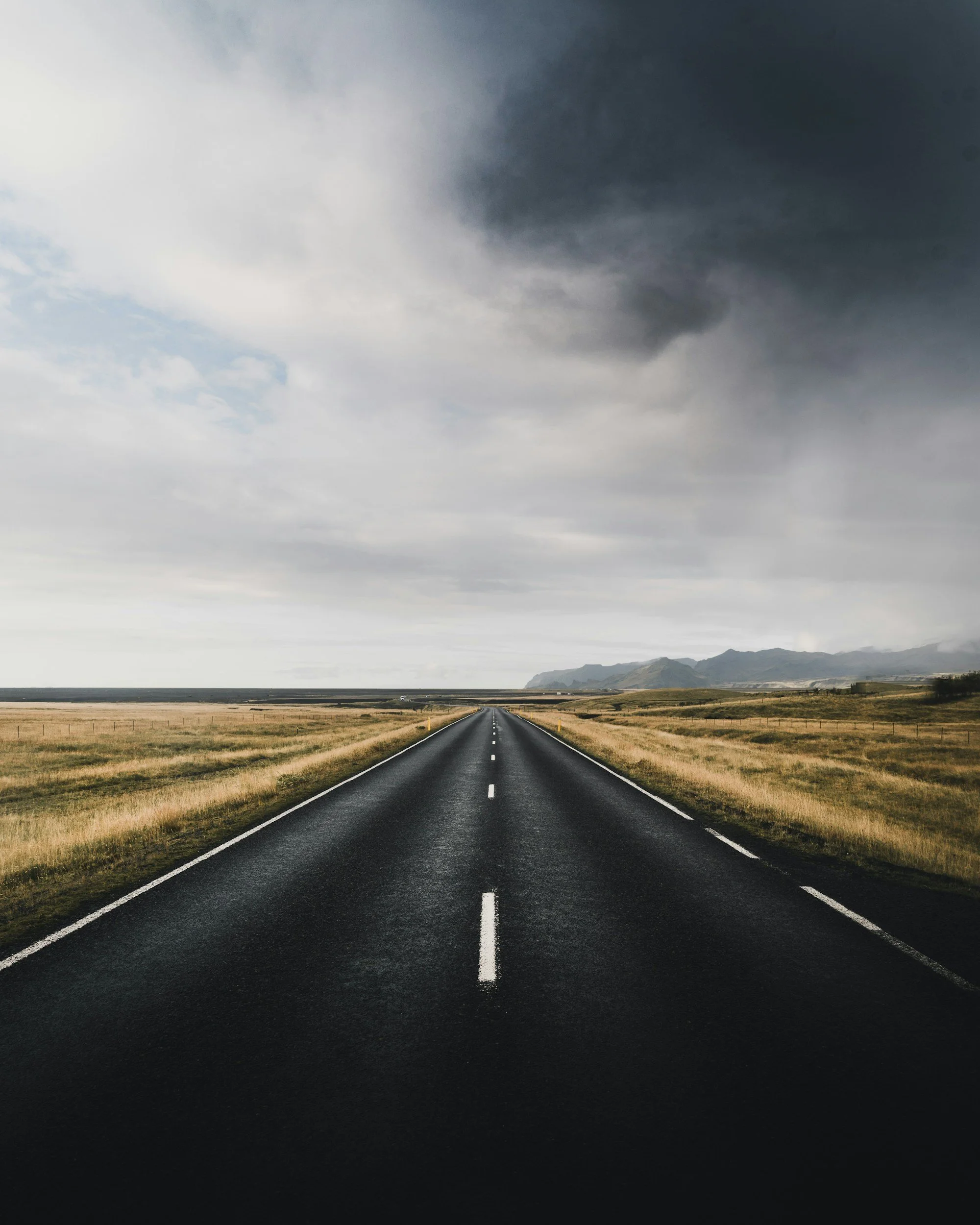 A straight, empty road running through a flat, grassy landscape under a cloudy, overcast sky.