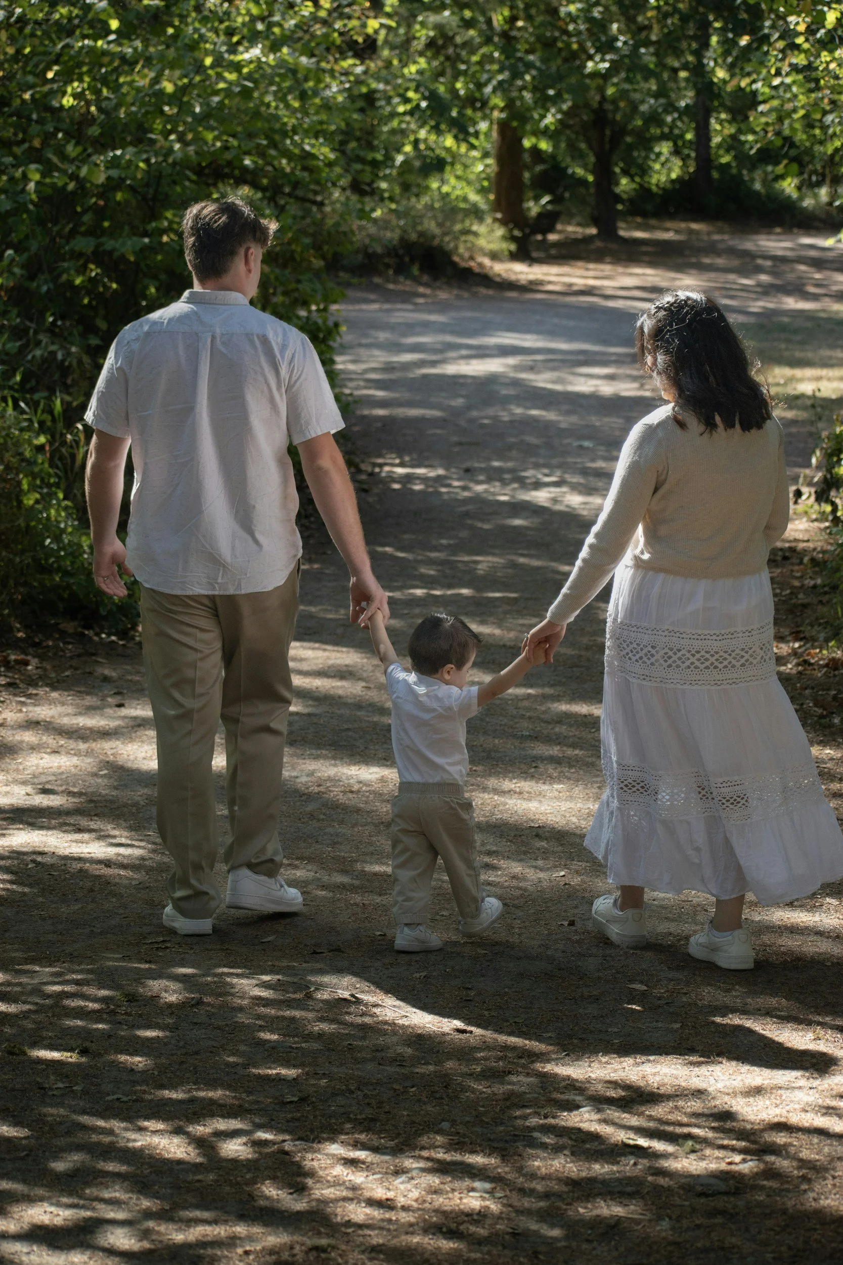 A family of three walking on a dirt path in a wooded park, holding hands.