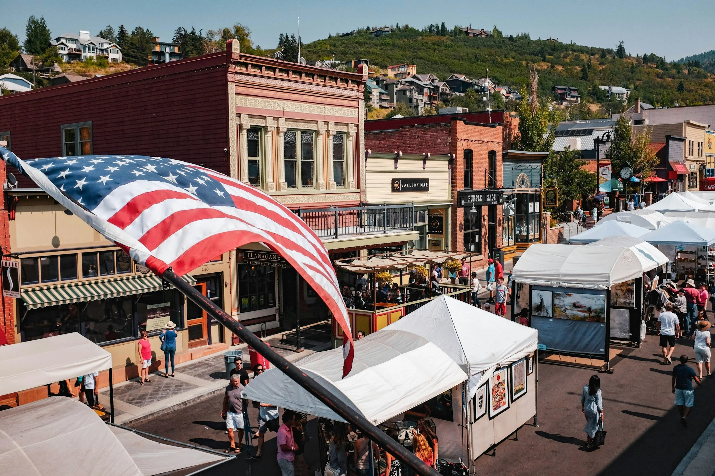 A small town Main Street with tents, storefronts, and pedestrians, featuring a waving American flag in the foreground.
