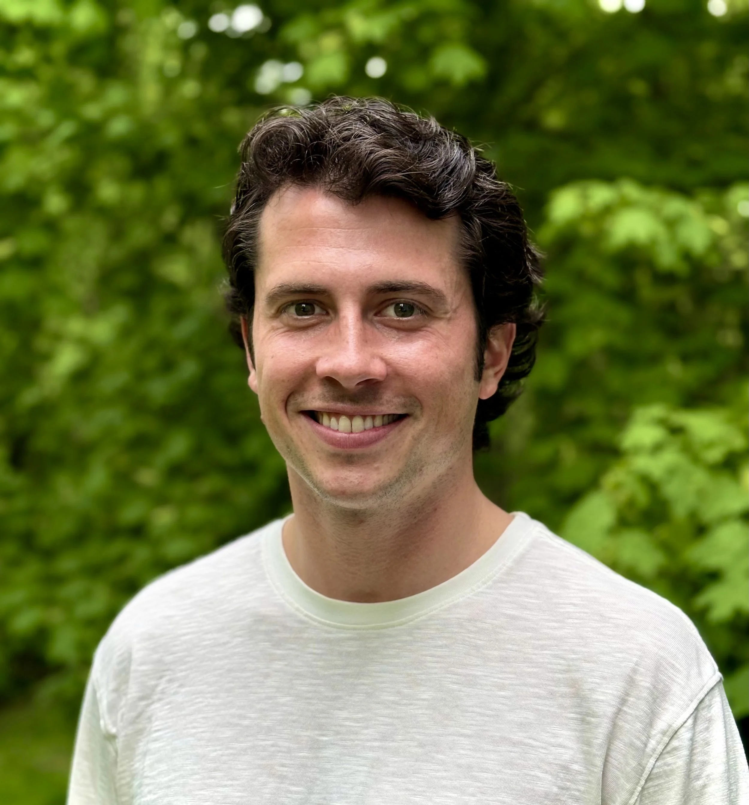 Smiling young man with dark, curly hair outdoors with green foliage in the background.