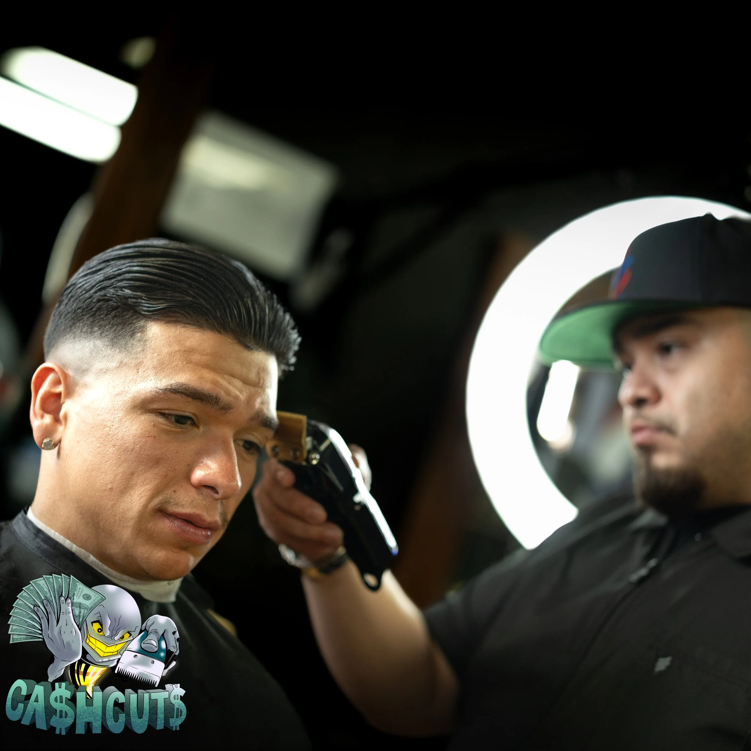 A man with slicked-back hair having his head shaved with electric clippers by another man wearing a cap, in a barber shop setting.