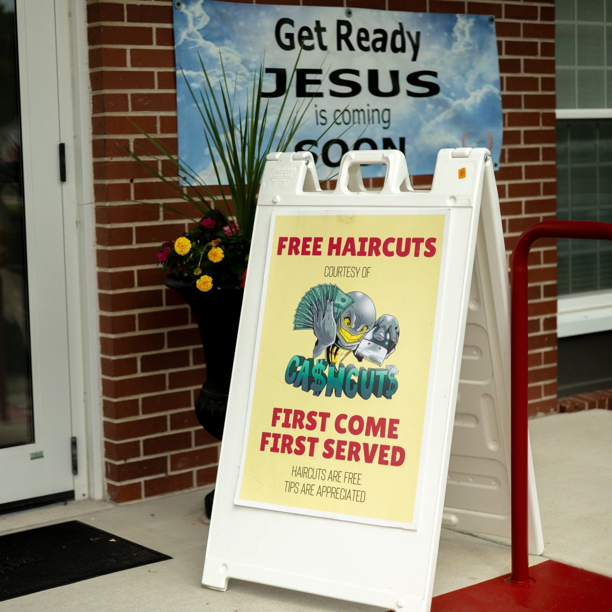 A sidewalk sign advertising free haircuts courtesy of Cash Cuts, with the slogan 'First Come First Served,' placed outside a building with a brick wall and a flower pot. A large banner in the background reads 'Get Ready Jesus is Coming Soon'.
