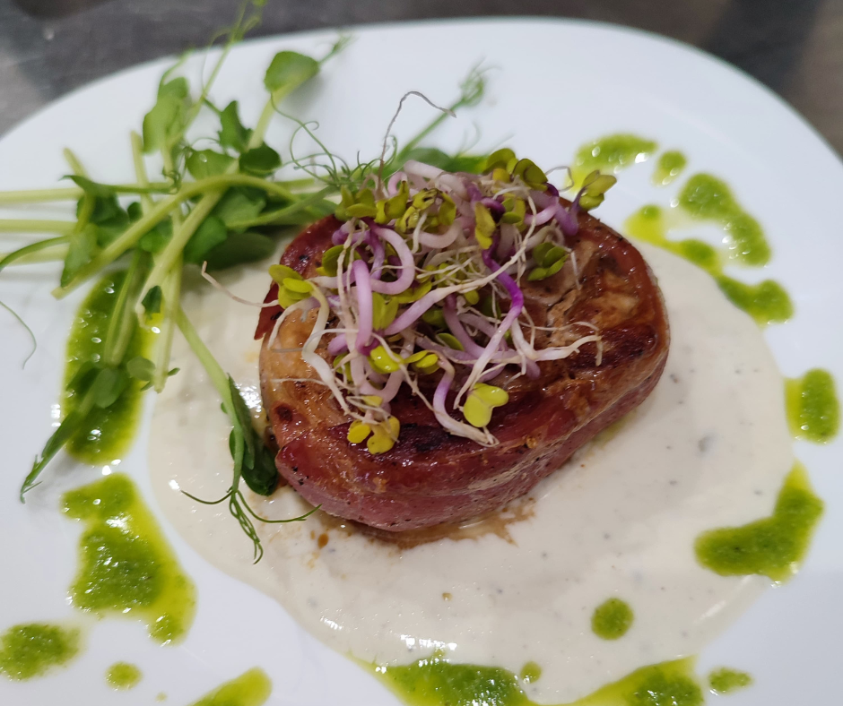 A steak topped with microgreens served with a white sauce and green sauce drizzled around on a white plate.