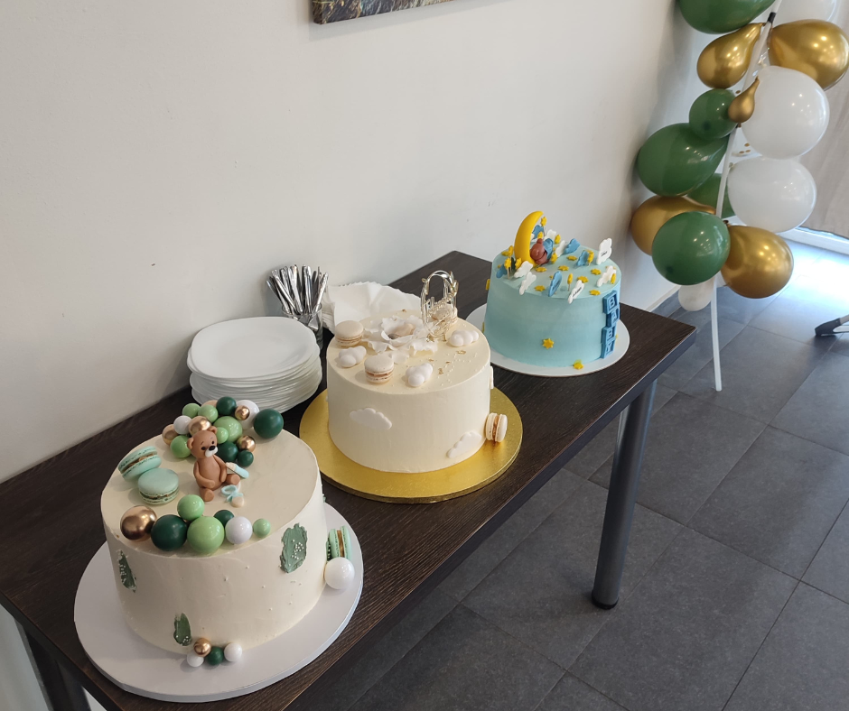 Three decorated cakes on a table, with a stack of plates, utensils, and balloons nearby, likely at a celebration.