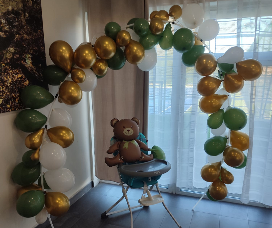 A teddy bear balloon decoration arch with white, green, and gold balloons surrounds a baby high chair with a teddy bear plush toy attached to it, in front of a window with curtains.